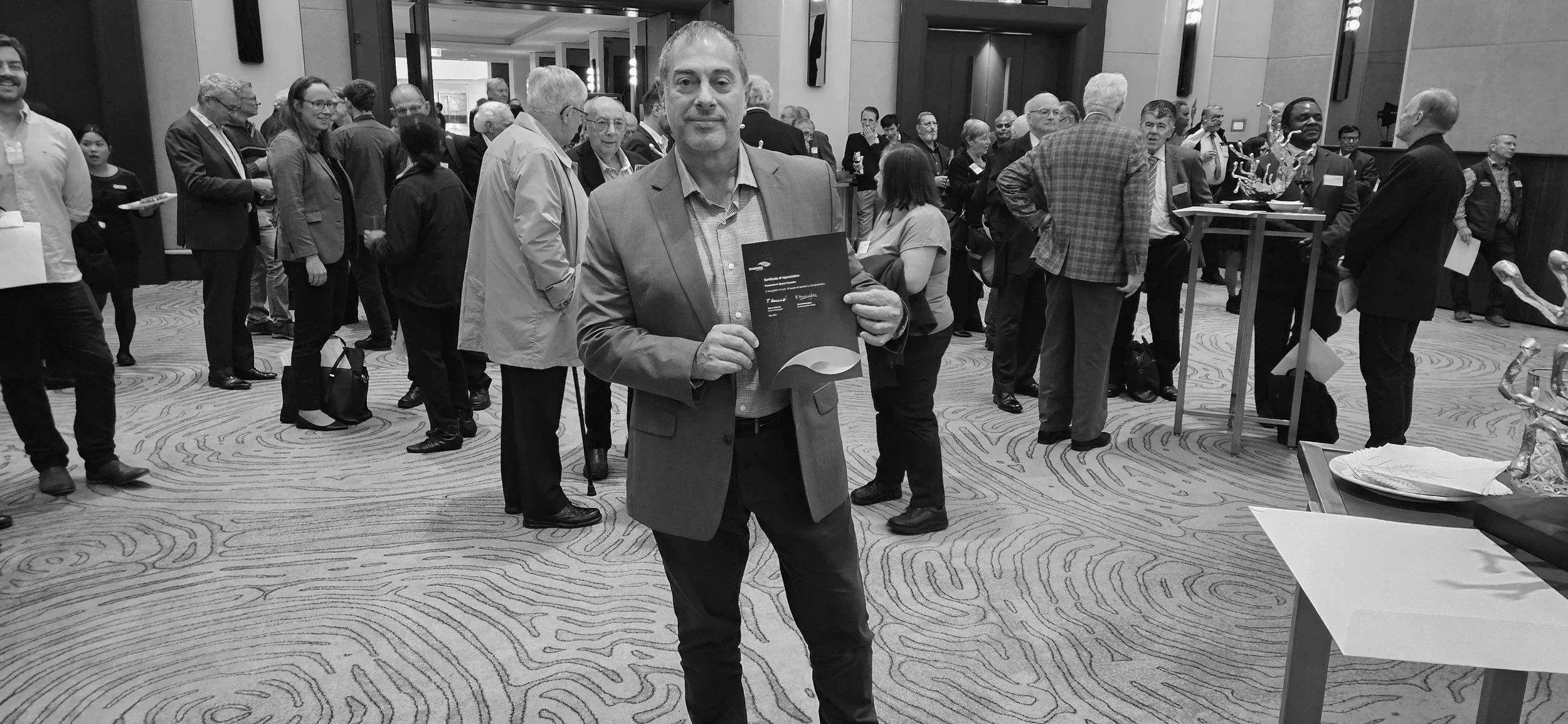 A man in a suit holding a certificate at a professional event, with a crowd of people mingling in the background.