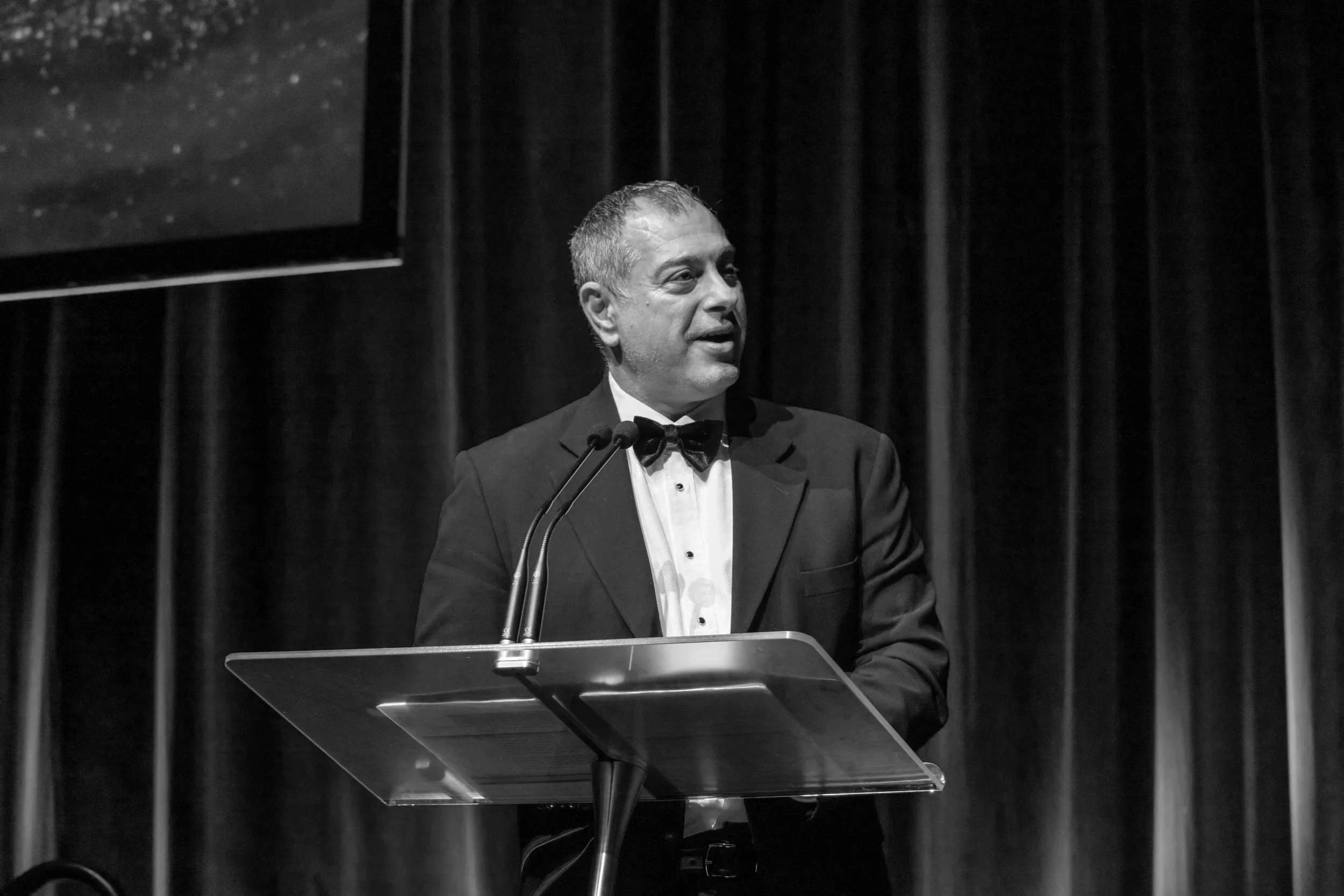 A man in a tuxedo and bow tie speaking at a podium with microphones, standing in front of a dark curtain.