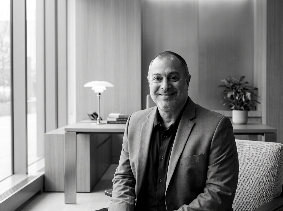 Black and white portrait of a smiling man in a suit sitting in an office with a desk, books, a table lamp, and potted plant.