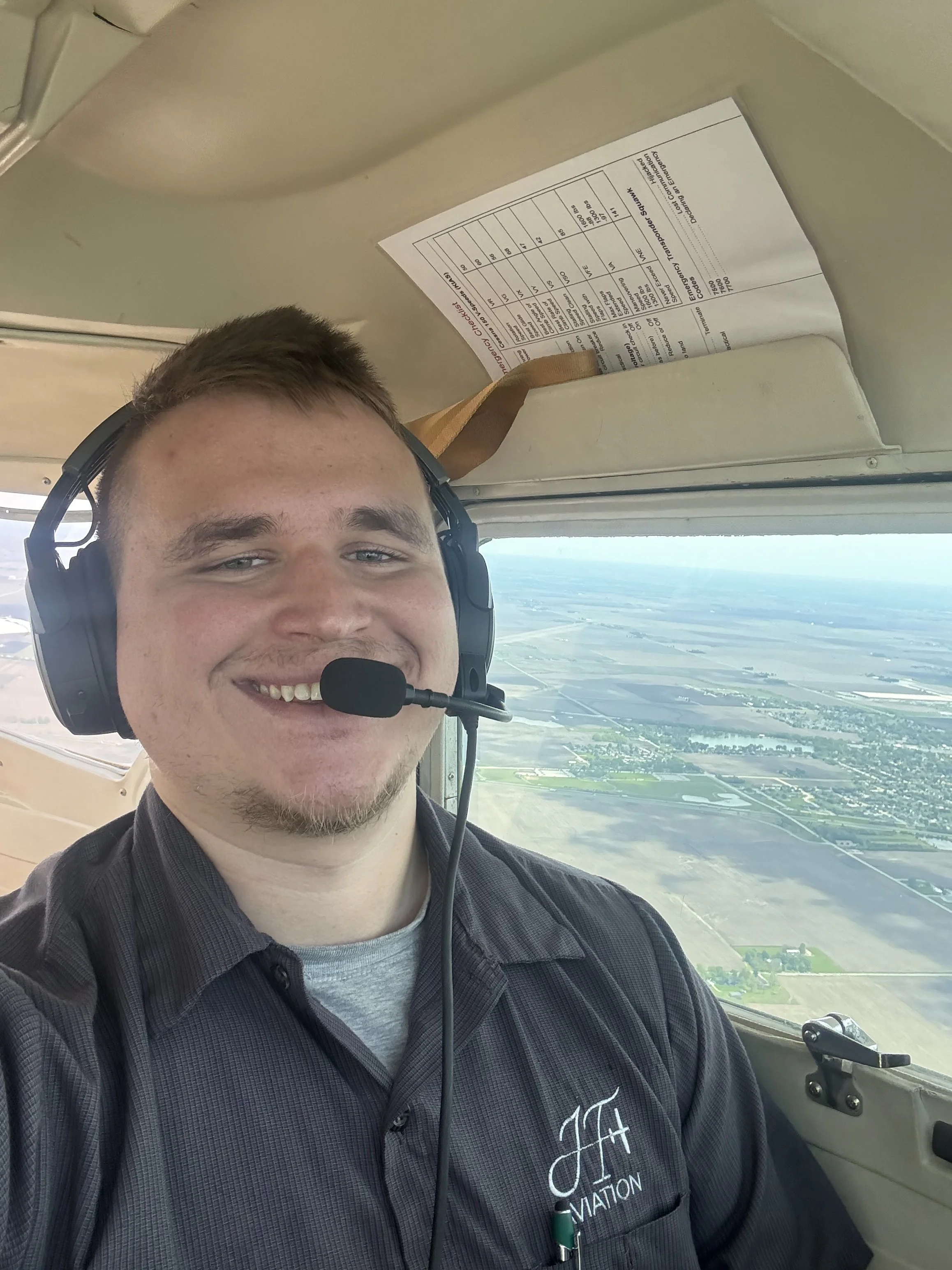 A young man smiling in the cockpit of an airplane, wearing a headset and a black shirt with a logo. The aircraft's window shows a view of fields and a river below.