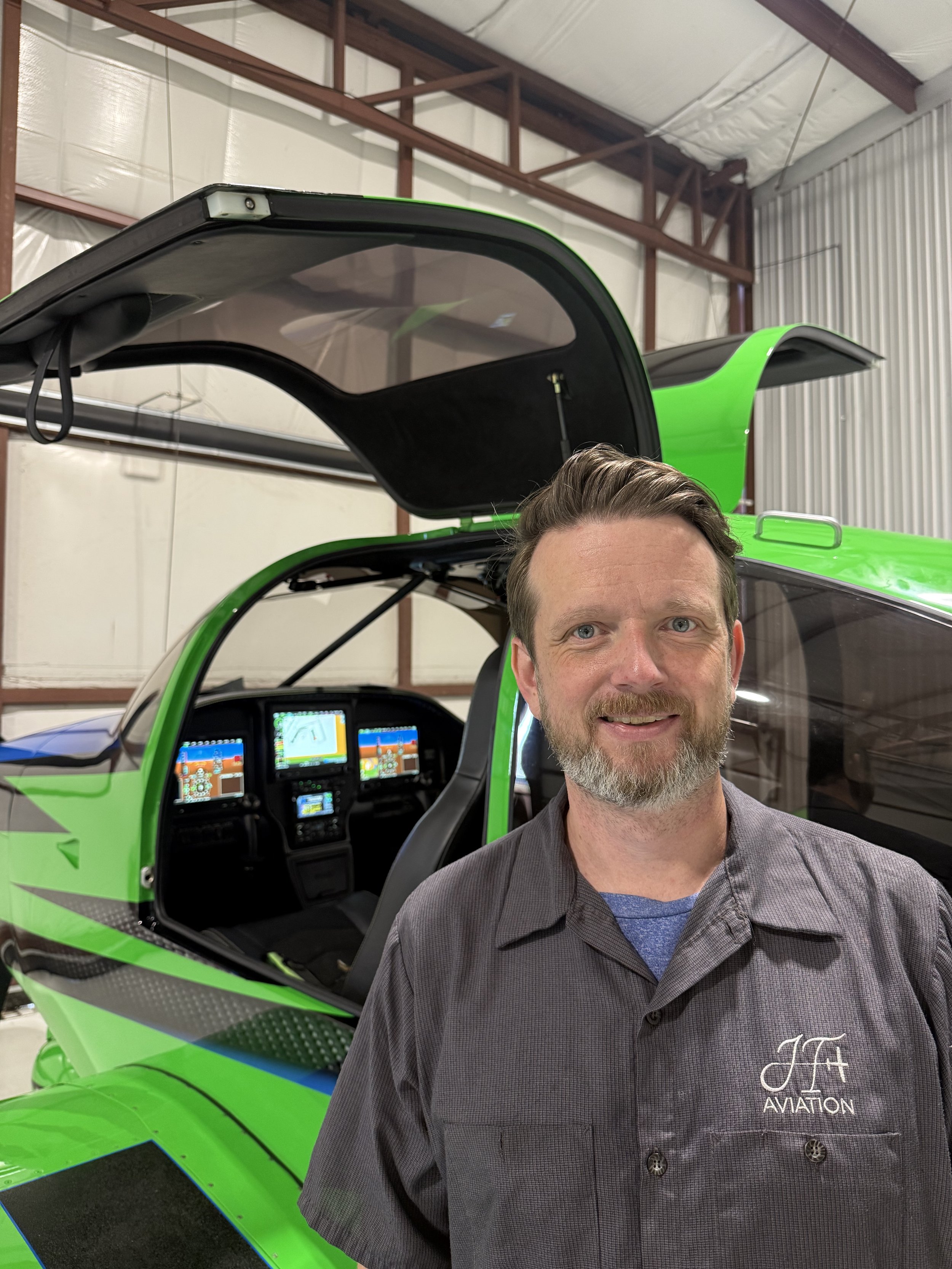 A man with a beard smiling standing in front of a bright green helicopter with its cockpit open inside a hangar.
