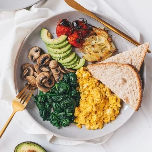 Breakfast plate with scrambled eggs, toasted bread, sautéed spinach, sliced avocado, sautéed mushrooms, roasted tomatoes, and hash browns, with a gold fork and knife.