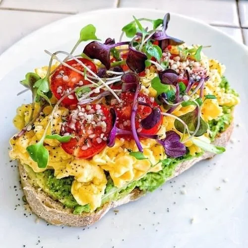 Open-faced sandwich on bread topped with scrambled eggs, cherry tomatoes, microgreens, and seasonings