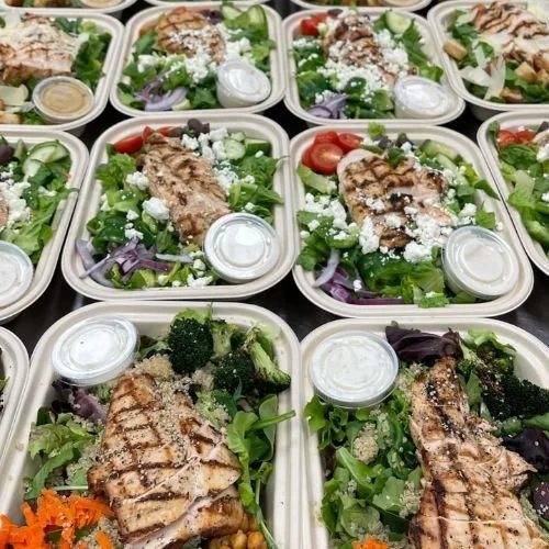 Multiple takeout salad containers filled with grilled chicken, mixed greens, cherry tomatoes, red onions, and small containers of dressing.