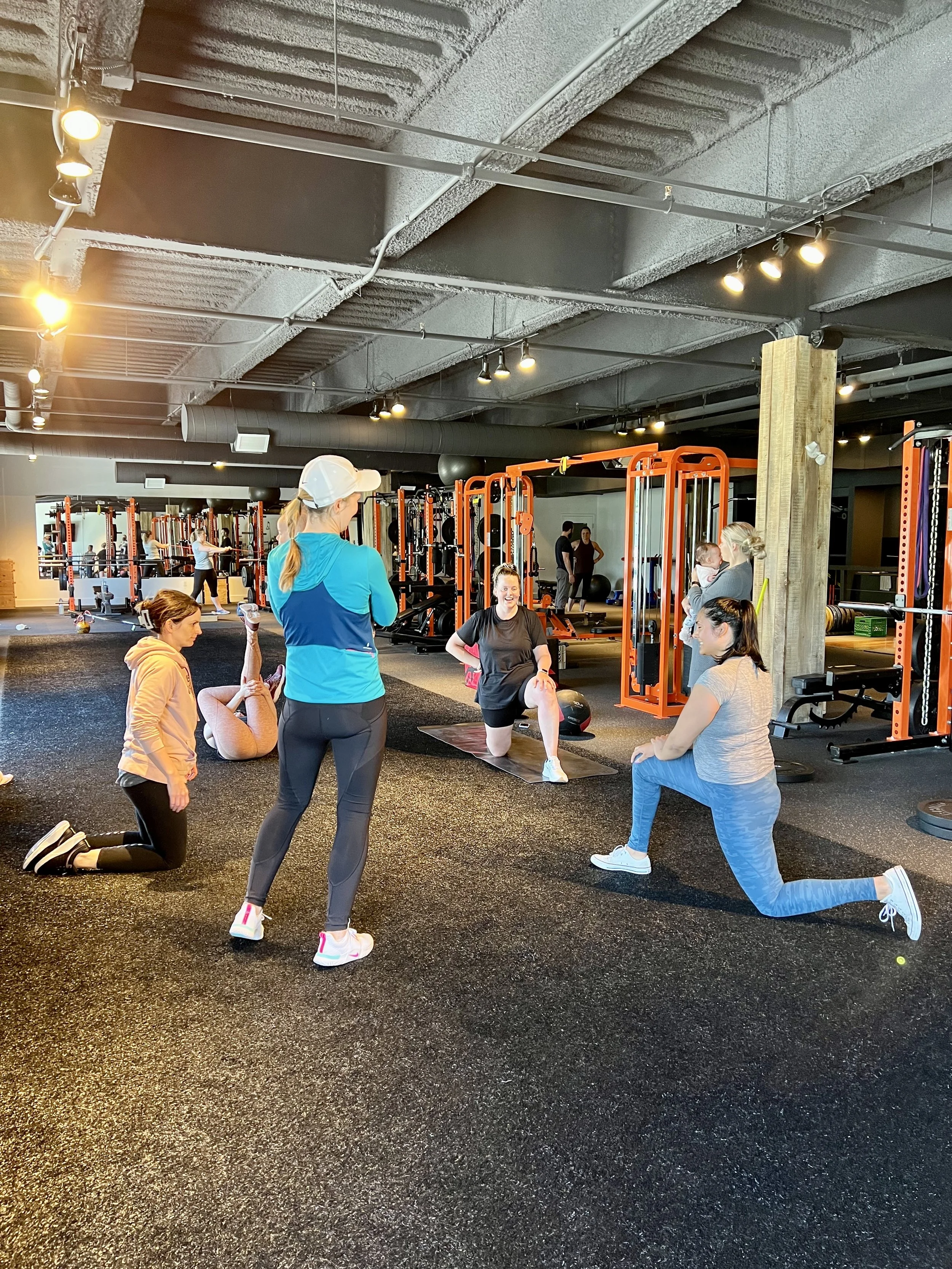 Group fitness class inside a gym, with women stretching, talking, and exercising on a black rubber floor. Gym equipment, including orange and black strength training machines and weights, is visible in the background.