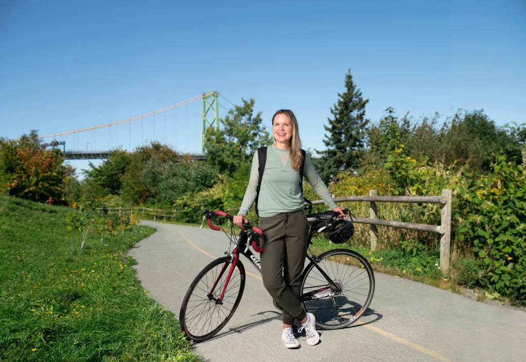 A woman standing with a bicycle on a paved trail, with a bridge and trees in the background under a clear blue sky.