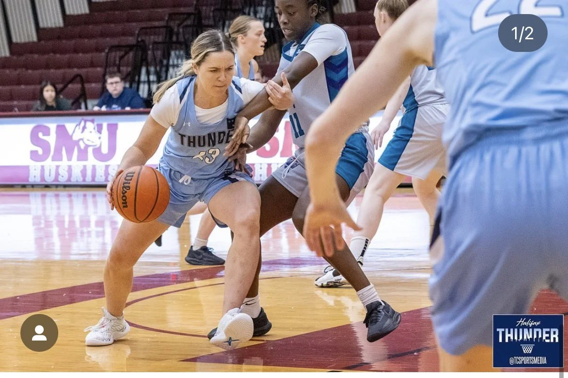 Women playing basketball on a court, one woman in a blue and white uniform dribbling the ball while being guarded by a player in a similar uniform