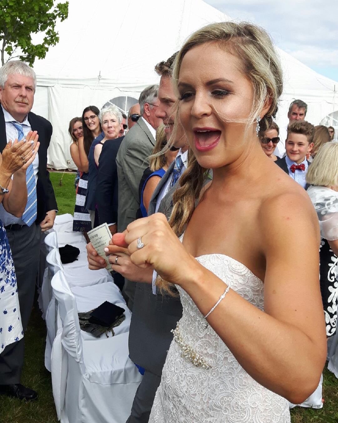 Bride in a white lace wedding dress with a braid hairstyle, surrounded by wedding guests at an outdoor reception under a large white tent.