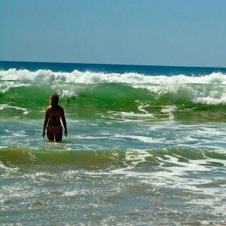 Woman standing in shallow ocean water facing green waves at the beach.