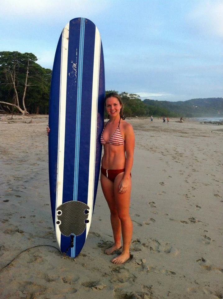 A woman in a red and white striped bikini stands barefoot on the sandy beach, smiling at the camera, next to a large blue and white stand-up paddleboard.