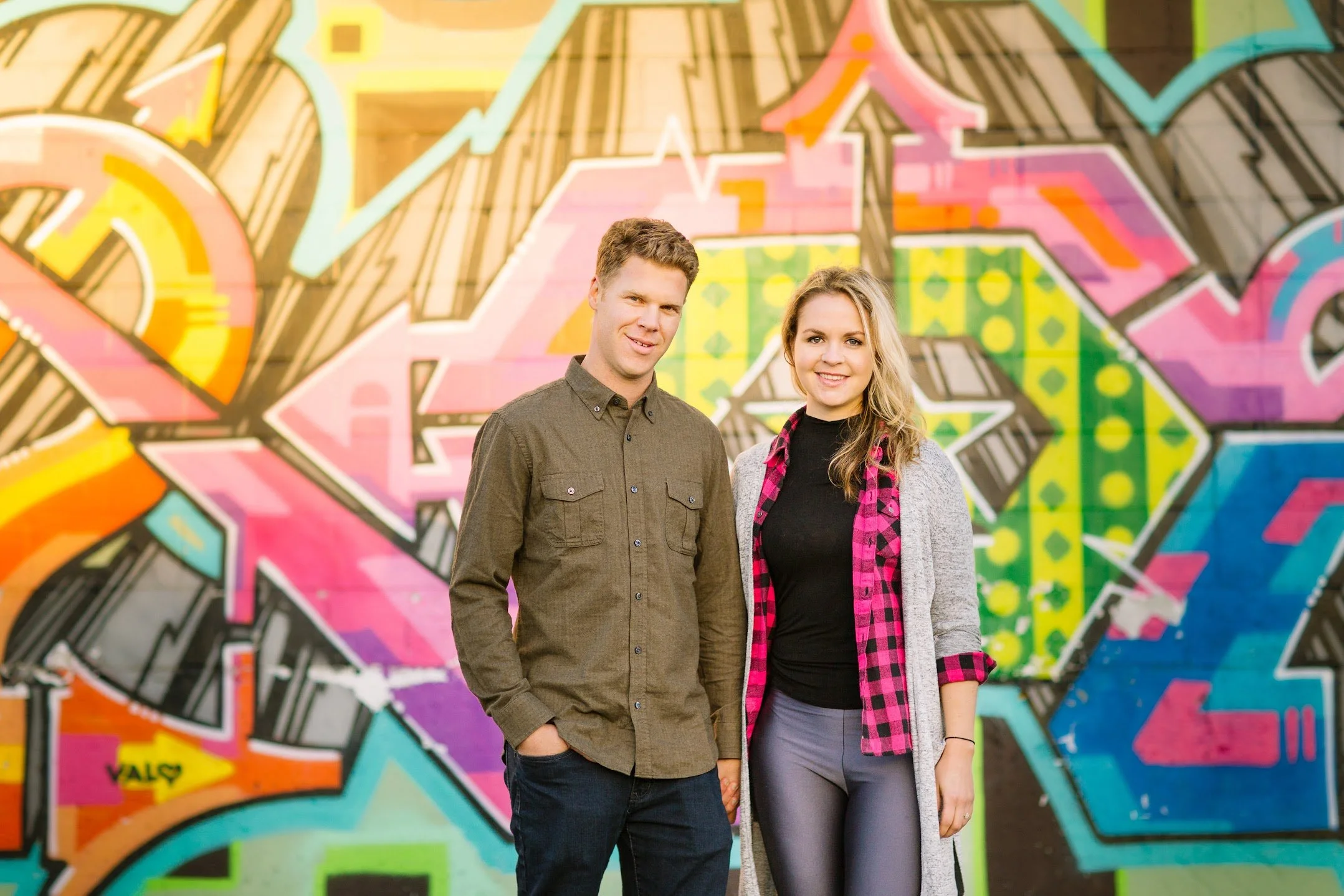 A young man and woman standing in front of a colorful graffiti wall.