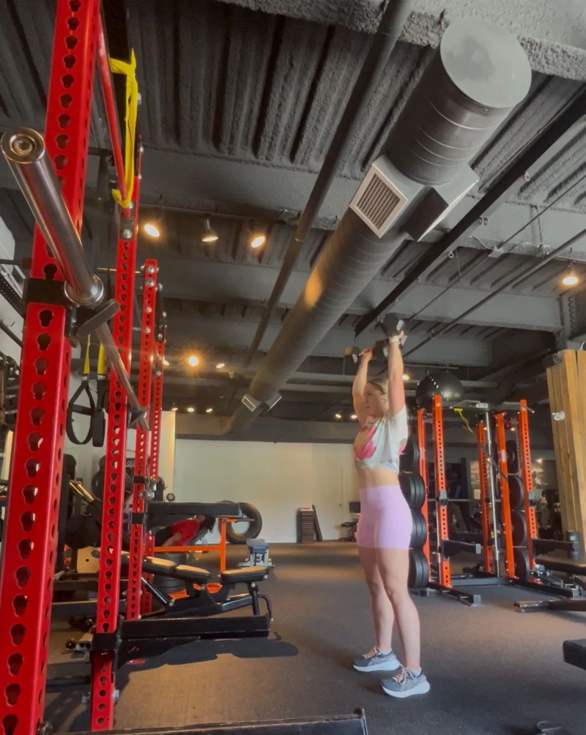 A woman lifting dumbbells overhead in a gym with orange and black workout equipment and a gray industrial ceiling.