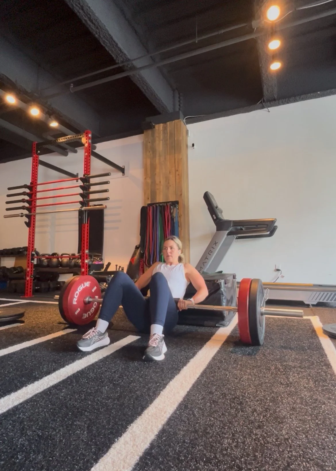 A woman in workout attire sitting on a gym floor, preparing to lift a barbell with red weight plates in a workout gym.