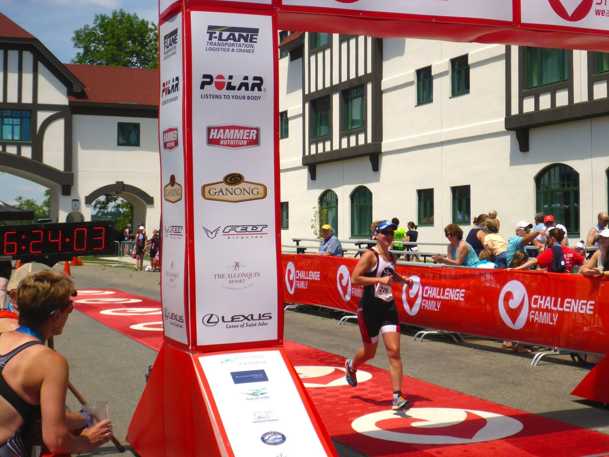 A woman running in a marathon, crossing the finish line with a crowd of spectators watching, race banners, and a digital clock displaying 6:24:03.