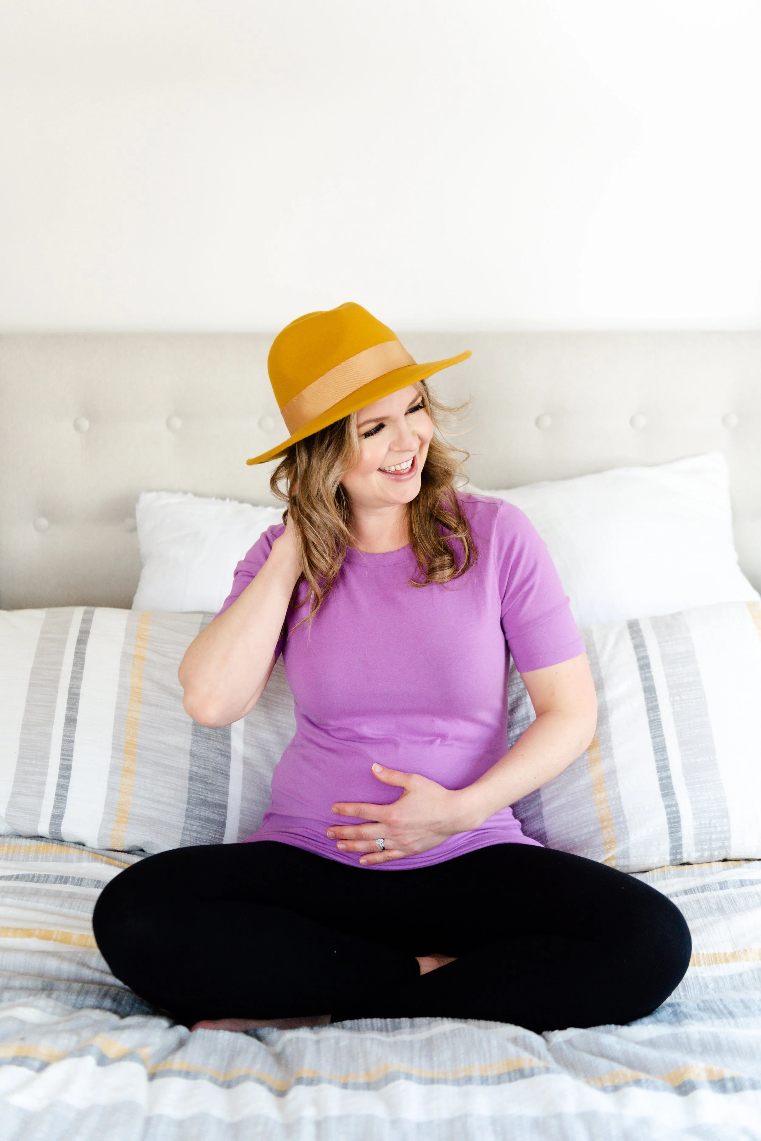 Woman with blonde hair wearing a yellow hat and a purple shirt, sitting cross-legged on a bed, smiling and touching her stomach.