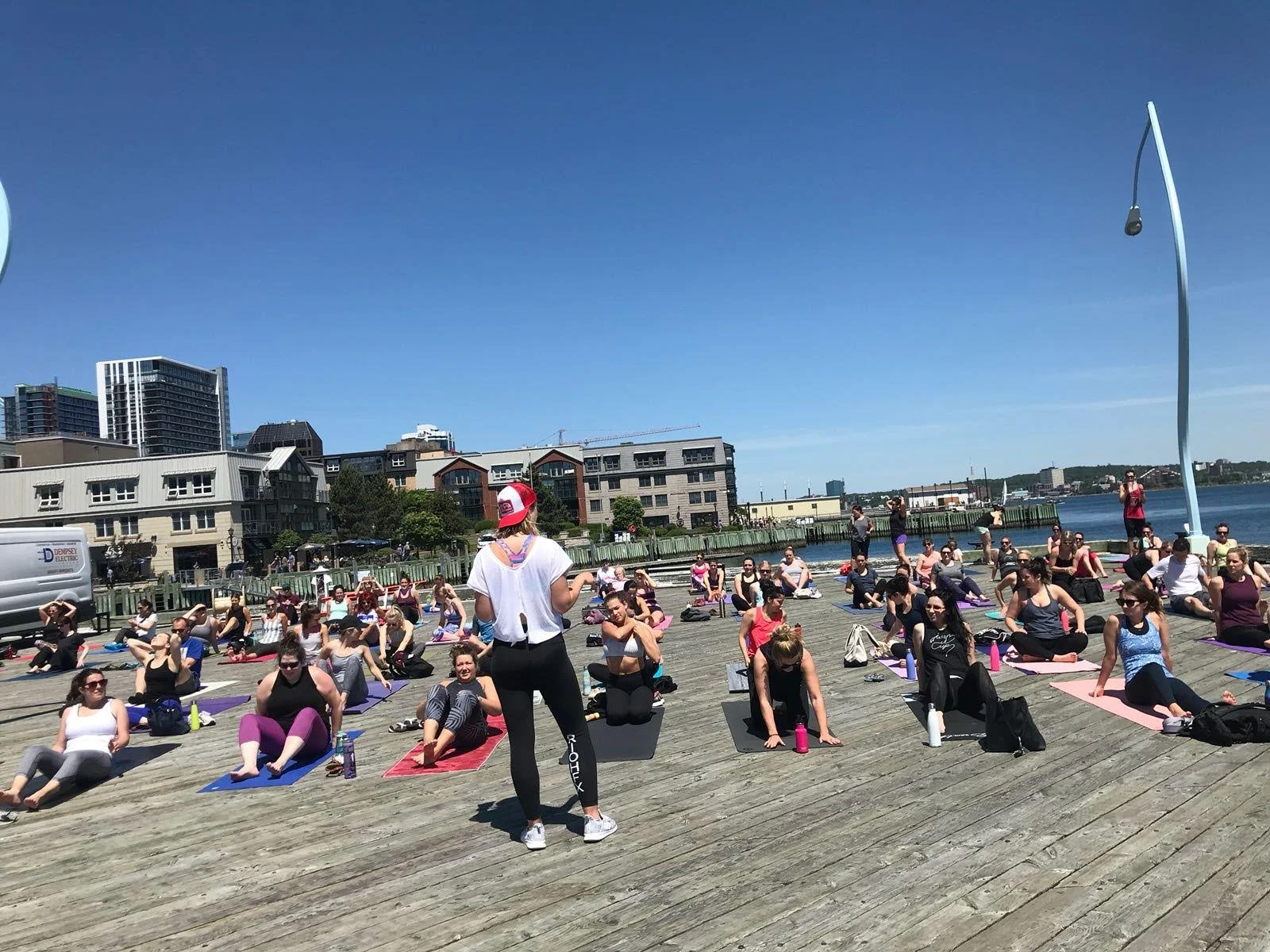A group of people participating in an outdoor yoga class on a wooden pier beside a body of water on a sunny day.