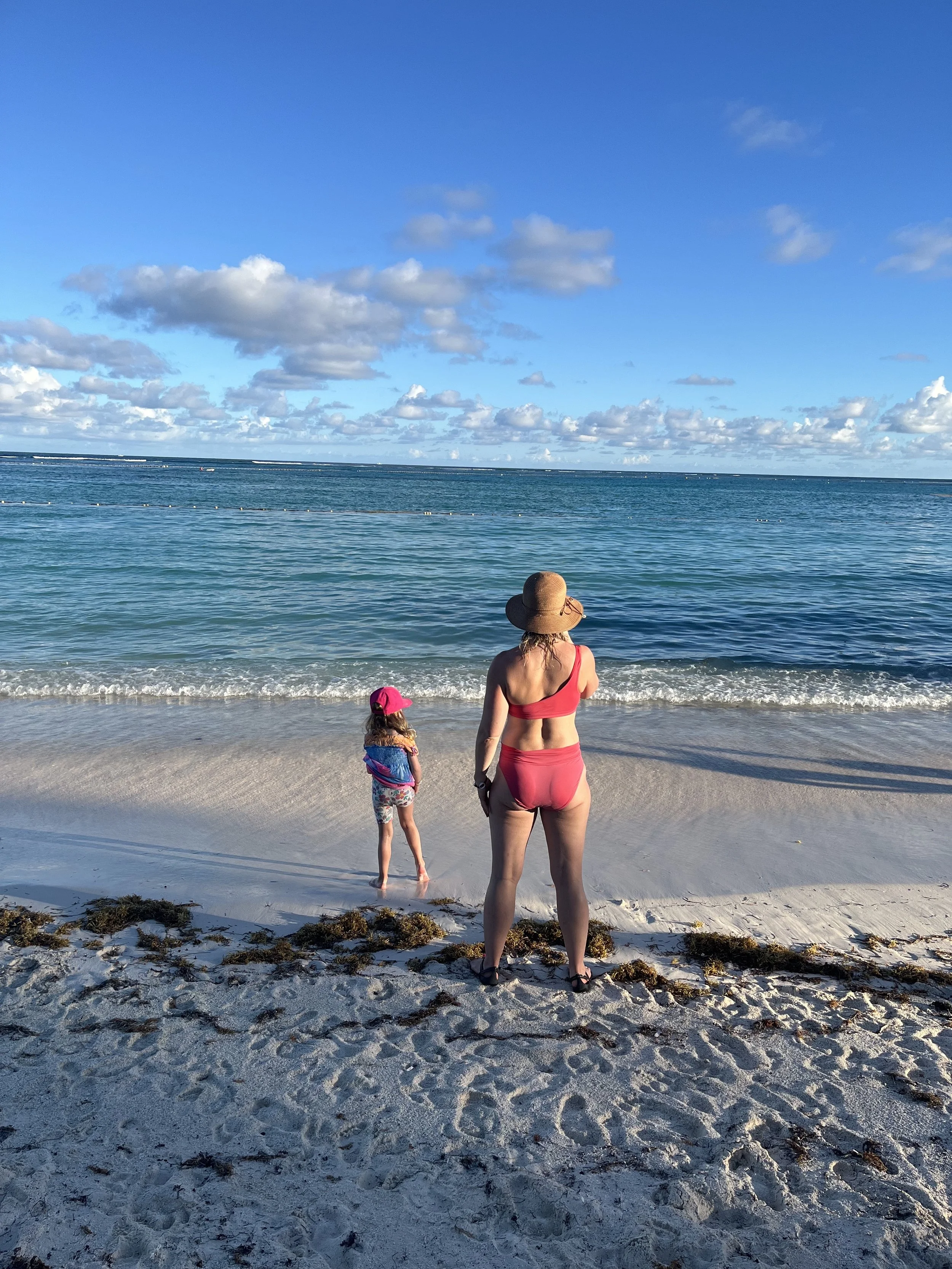 Woman in a red swimsuit and wide-brimmed hat standing on a sandy beach near a child in a pink hat and colorful swimsuit, with the ocean and blue sky with clouds in the background.