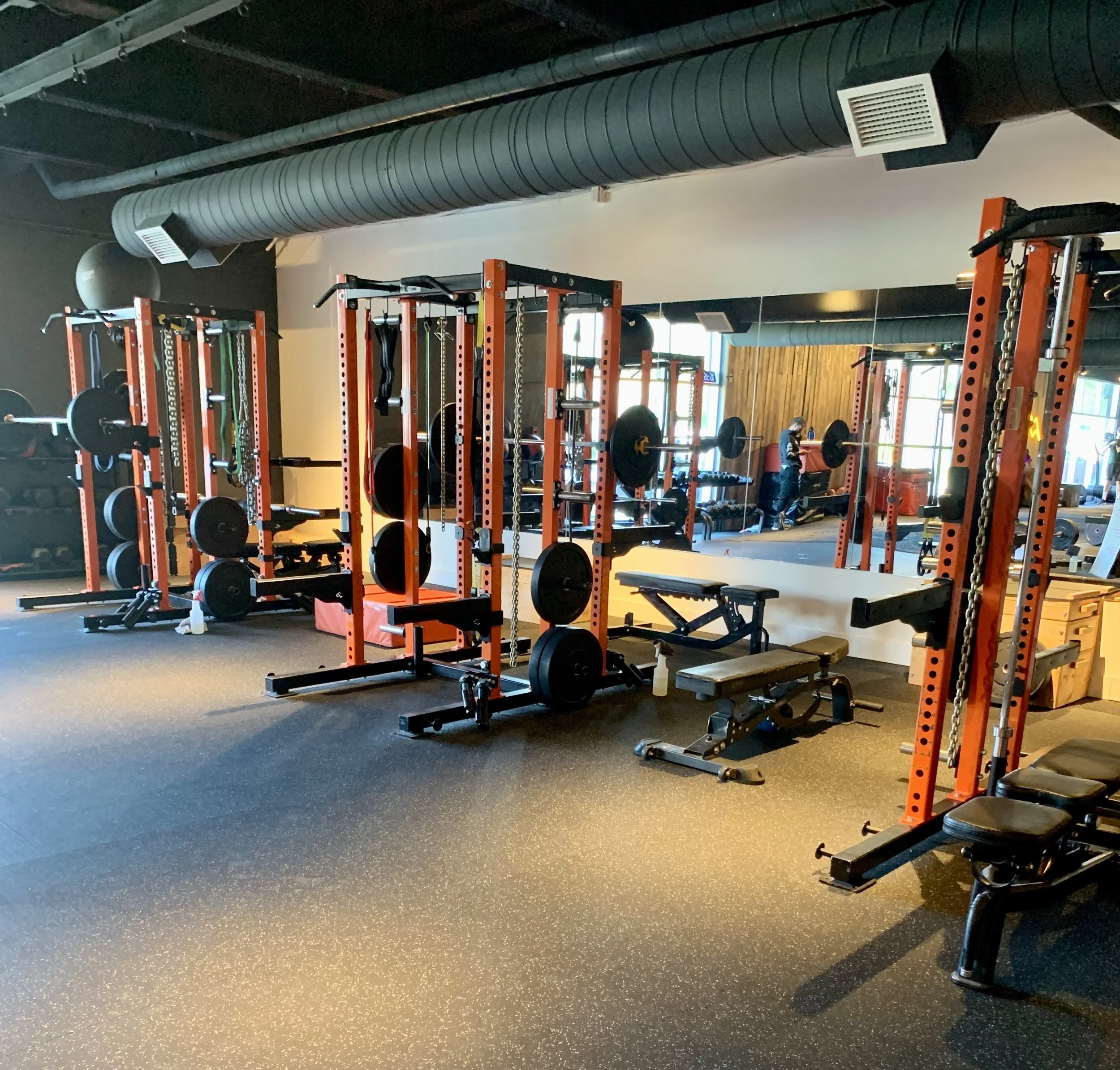 Interior of a gym with orange power racks, weight benches, and various weights. Large windows and a mirrored wall reflect exercise equipment.