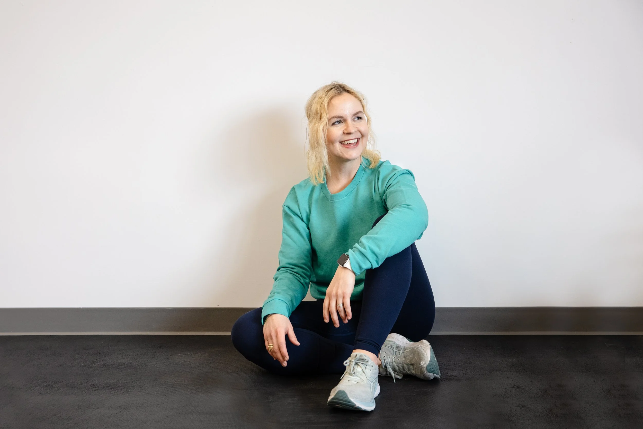 A smiling woman with blond hair sitting on a black floor against a plain white wall, wearing a teal sweatshirt, dark leggings, and running shoes, with an Apple Watch on her wrist.