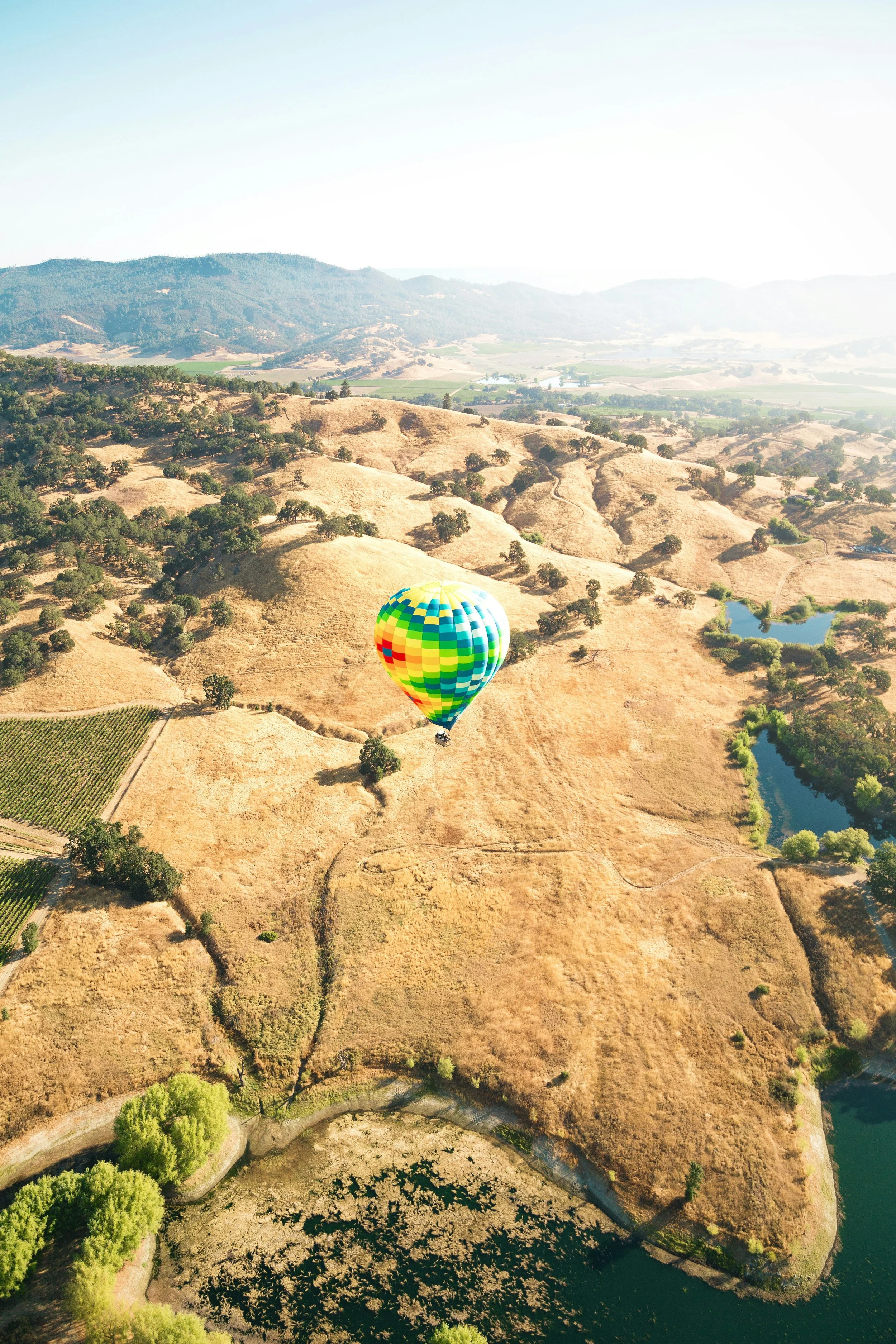 A rainbow-colored hot air balloon flying over dry, hilly landscape with patches of trees, water bodies, and farmland. Napa Valley Balloon ride