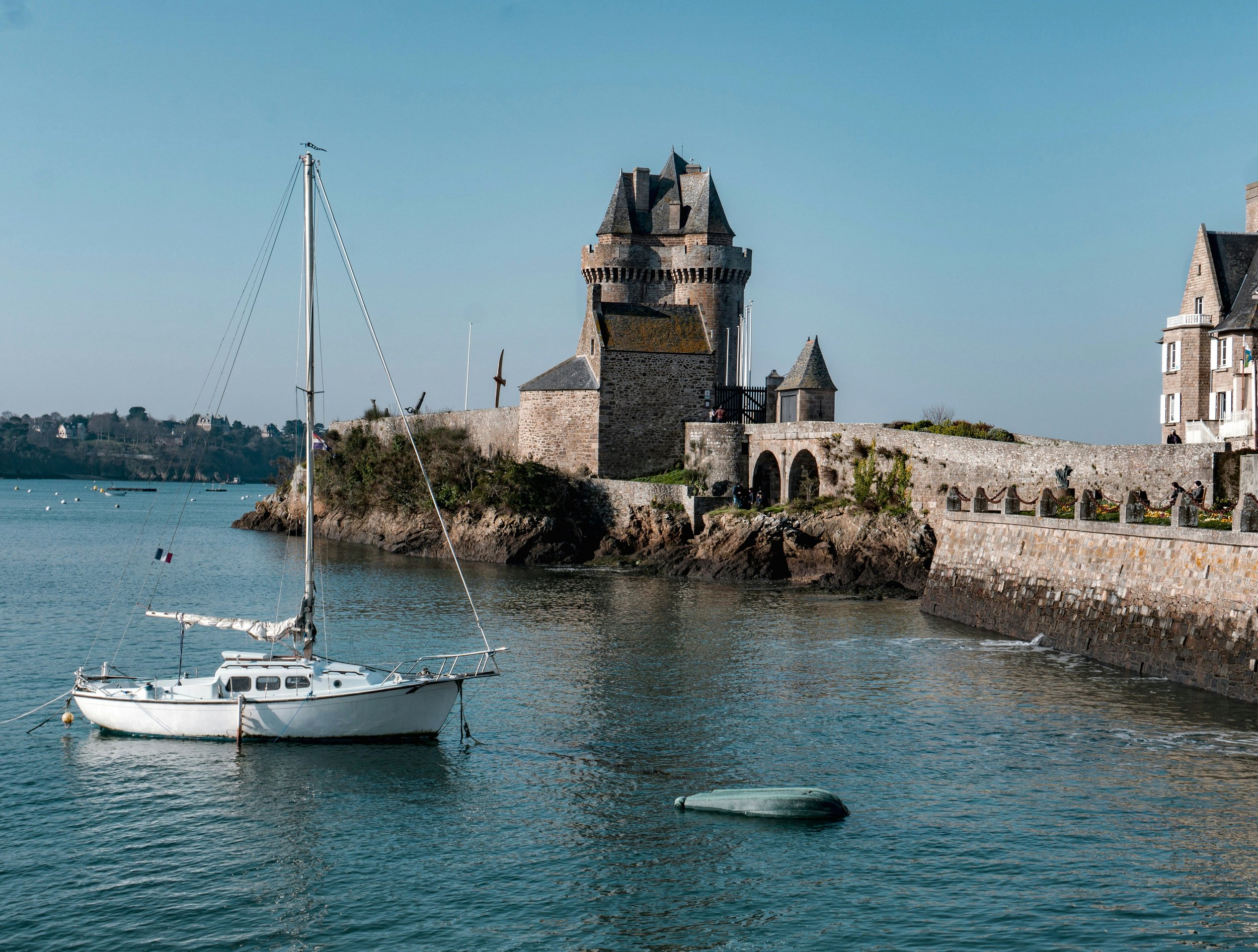 A white sailboat anchored in a calm harbor with a historic stone castle and stone wall along the shoreline, under a clear blue sky. VOYA curates bespoke travel experiences in France and Napa Valley.