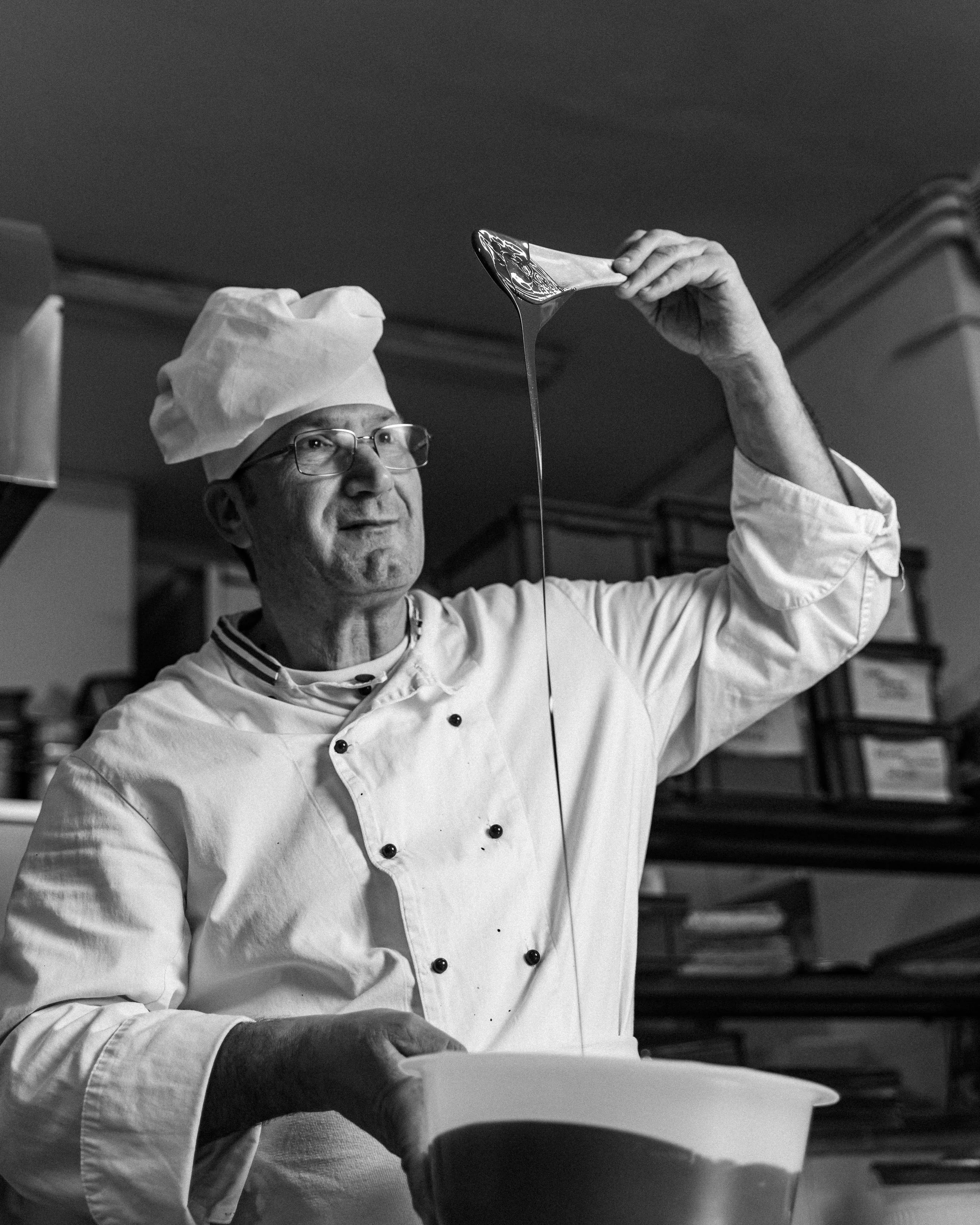 A male chef in a chef's coat and hat pouring batter from a large spoon into a bowl in a kitchen.