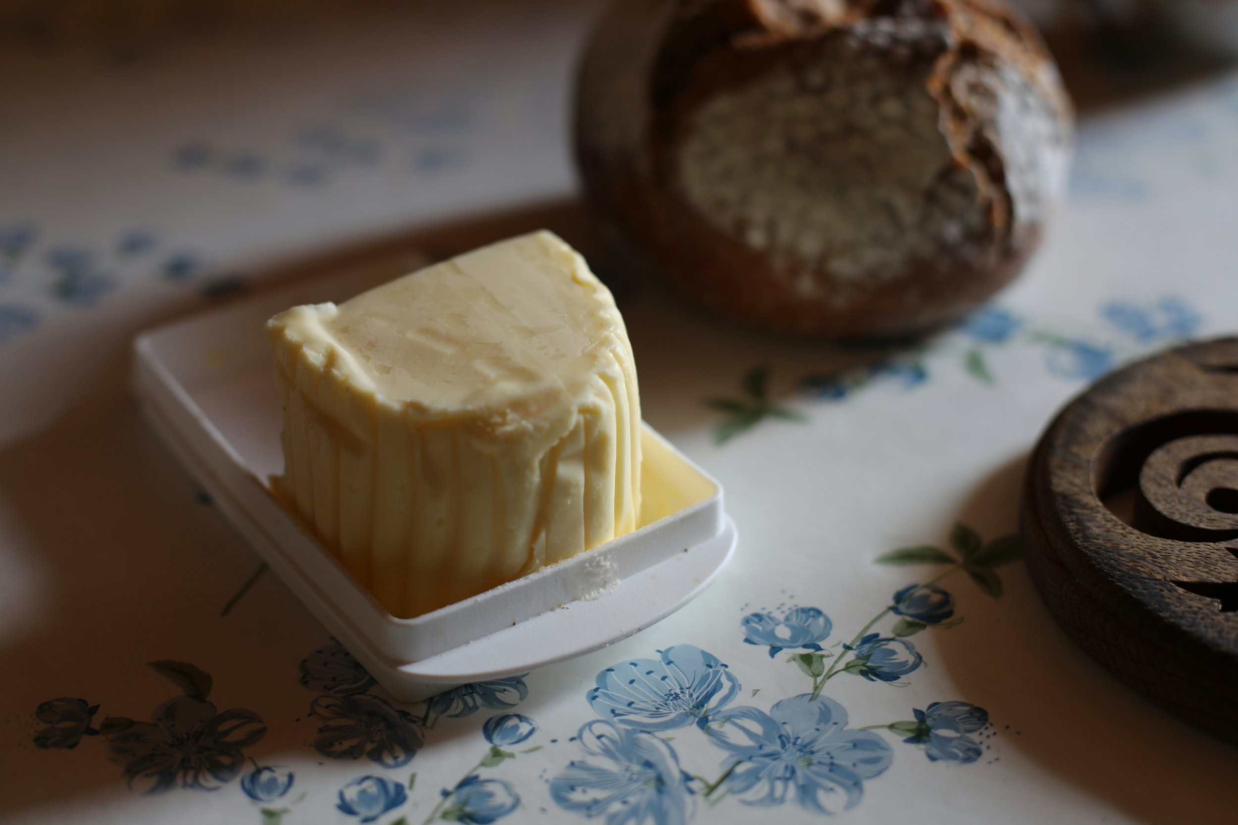 A block of butter in a butter dish on a floral tablecloth with whole bread and a round wooden object in the background. VOYA curates bespoke travel experiences in France and Napa Valley.