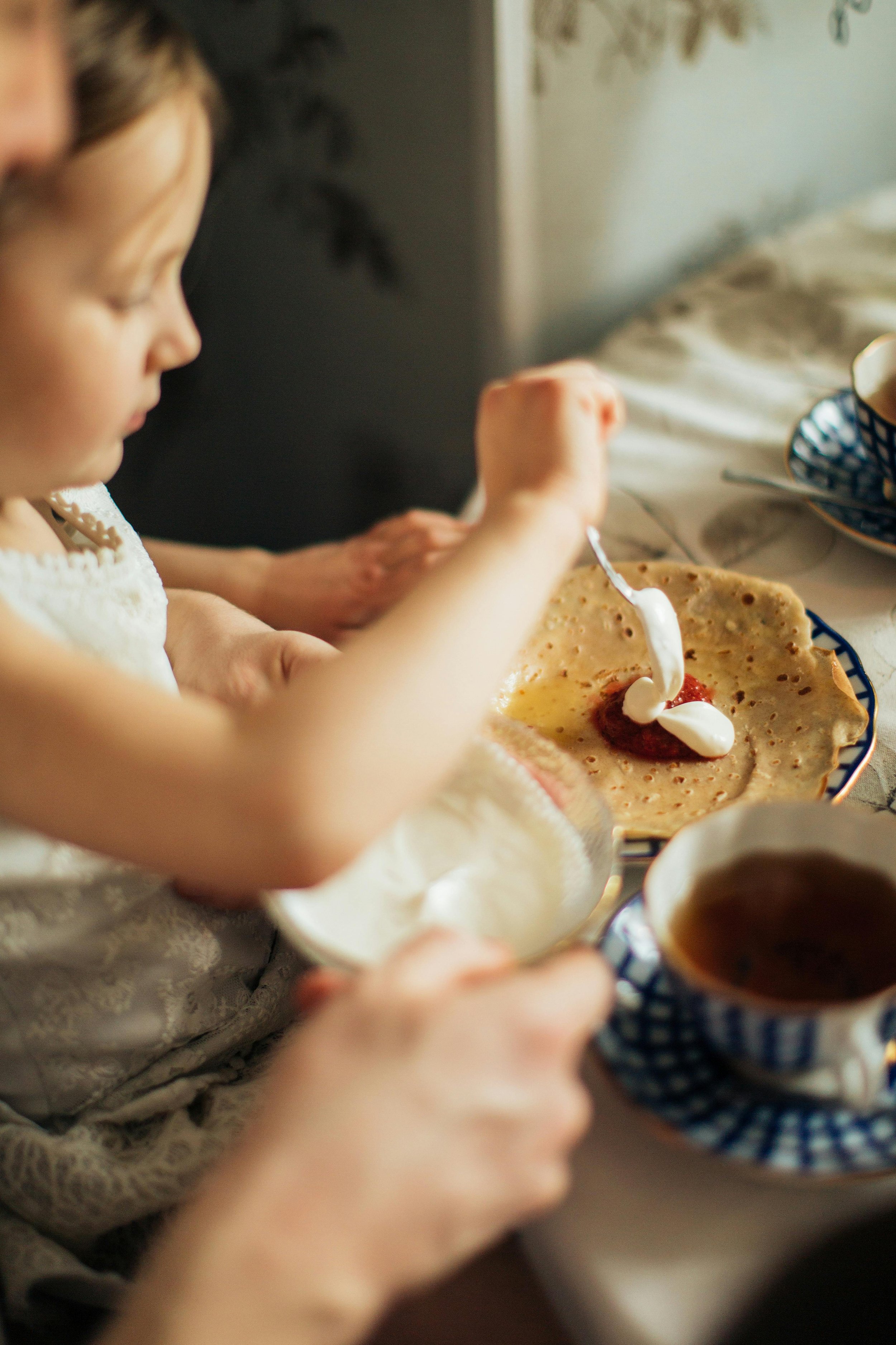 Close-up of a young girl spreading whipped cream onto a crepe with strawberries, sitting at a table with a cup of tea, in a cozy setting. VOYA curates bespoke travel experiences in France and Napa Valley.