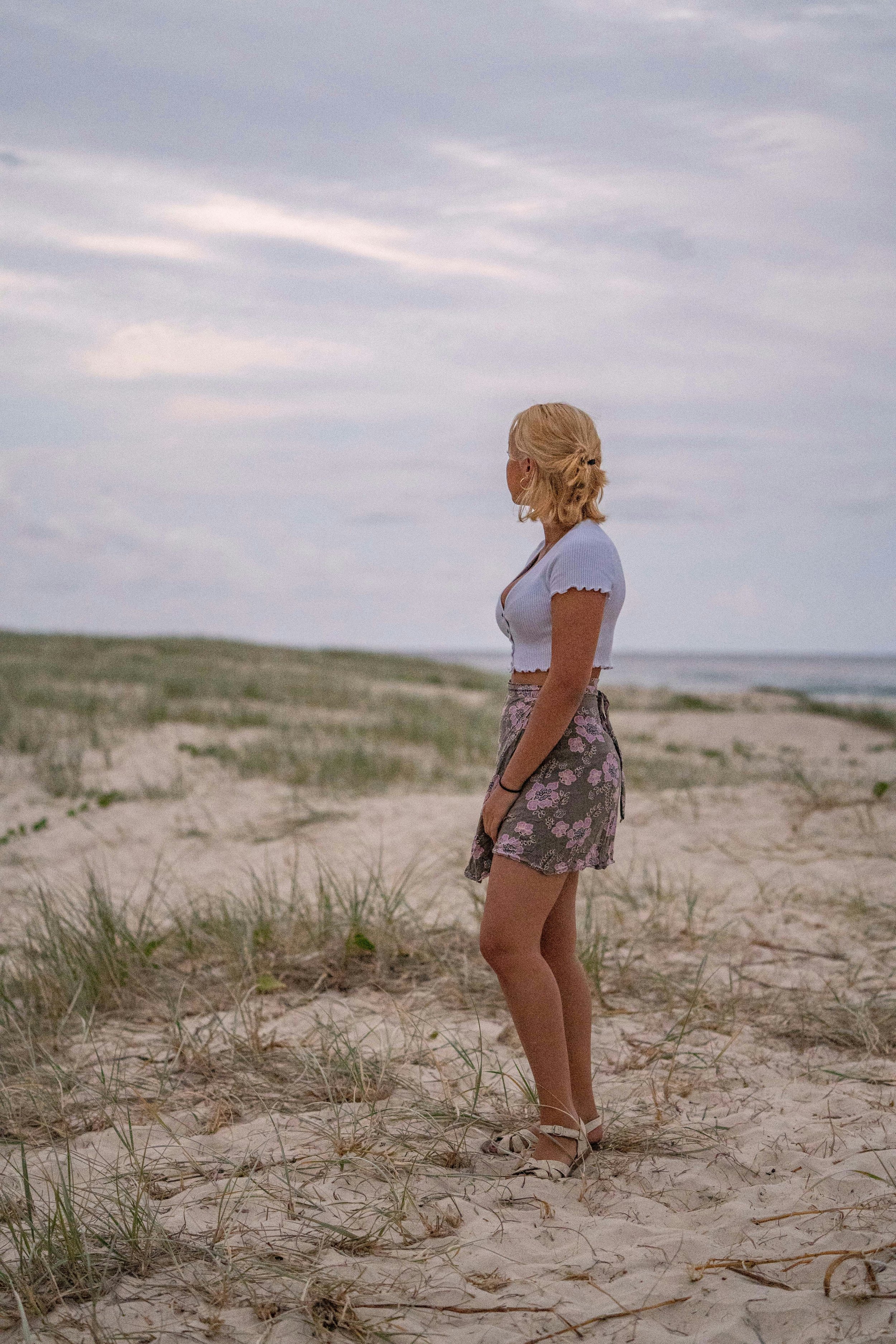 A woman with blonde hair in a braid, wearing sunglasses, a white short-sleeve crop top, floral shorts, and sandals, standing on a sandy beach with grass, facing the ocean under a cloudy sky.