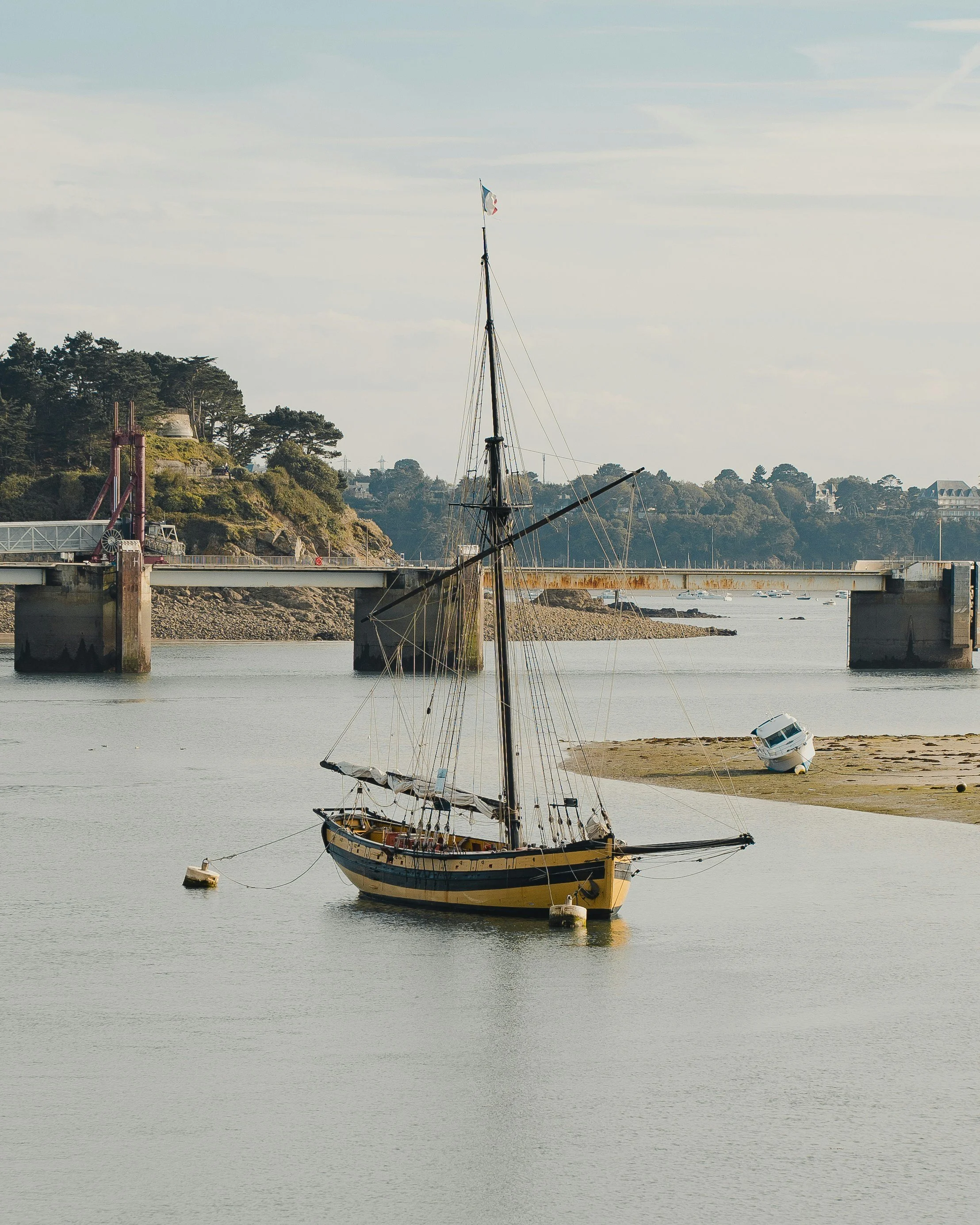A sailboat anchored in calm water near a sandy shoreline with a small boat overturned in the background, and a bridge with supporting pillars stretching across the water with trees and buildings in the distance.