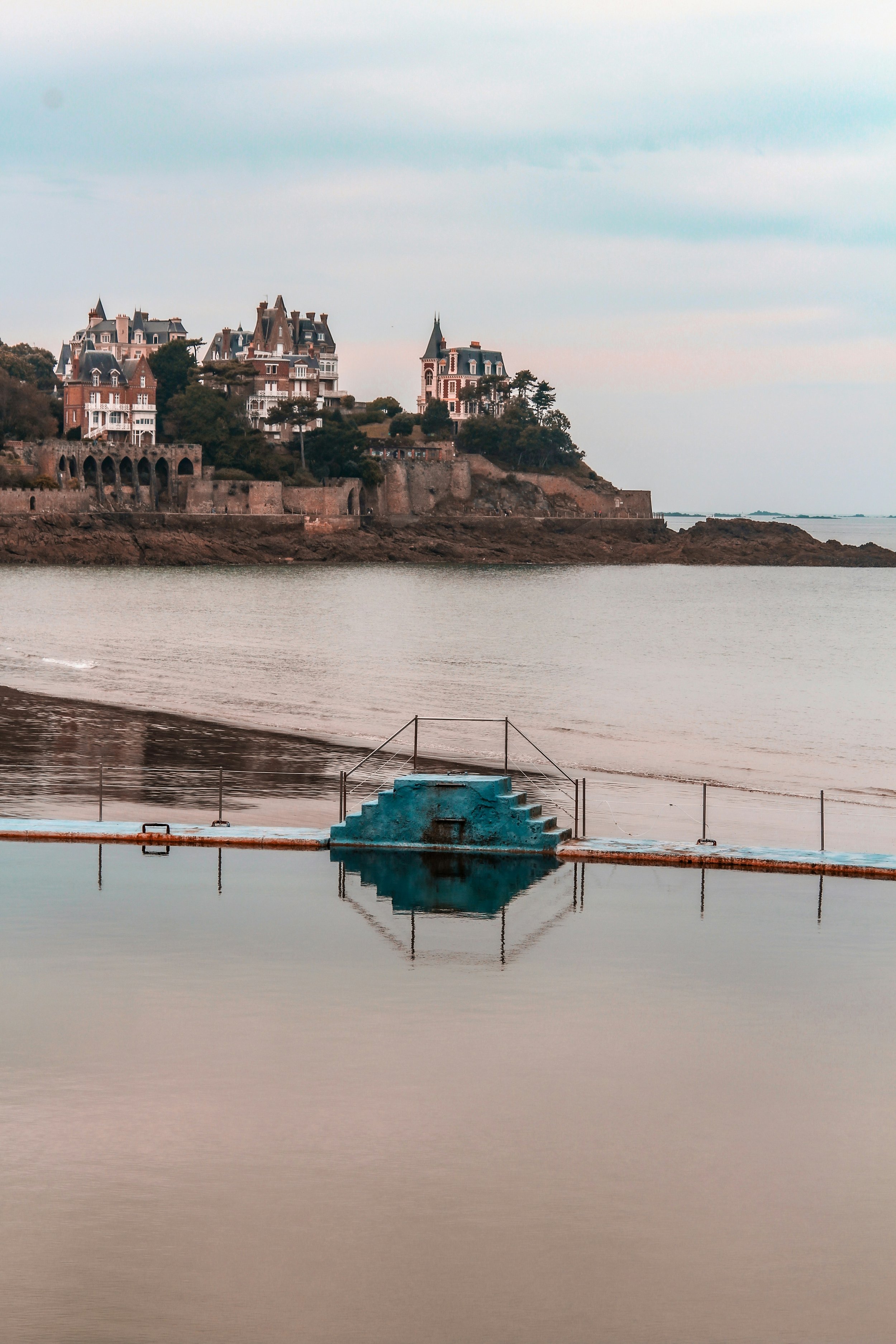 A calm body of water with stairs leading into it, with a European-style castle-like building on a hill in the background surrounded by trees, VOYA curates bespoke travel experiences in France and Napa Valley.