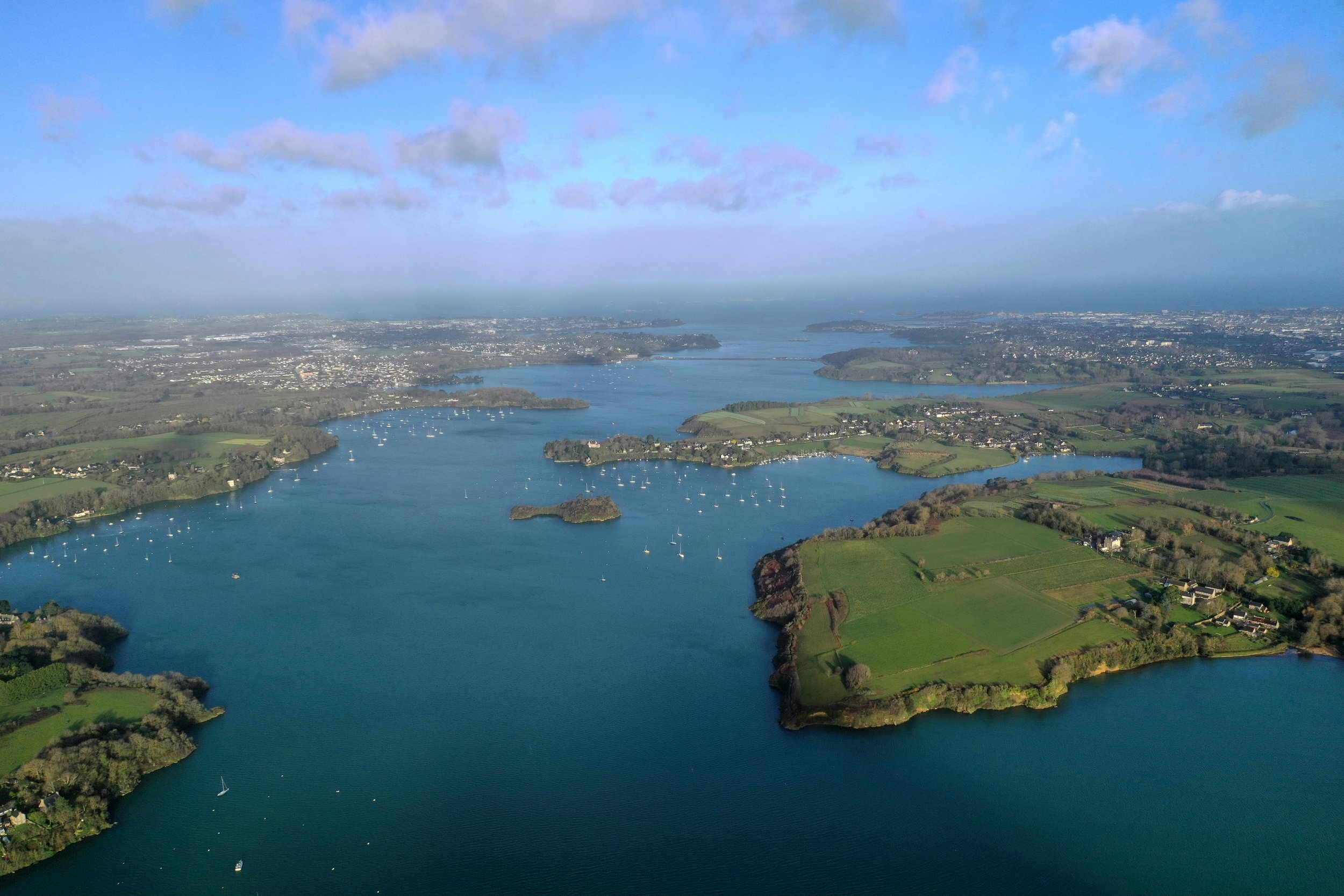 Aerial view of a large lake with several small islands, surrounded by green fields, houses, and a distant cityscape under a partly cloudy sky.