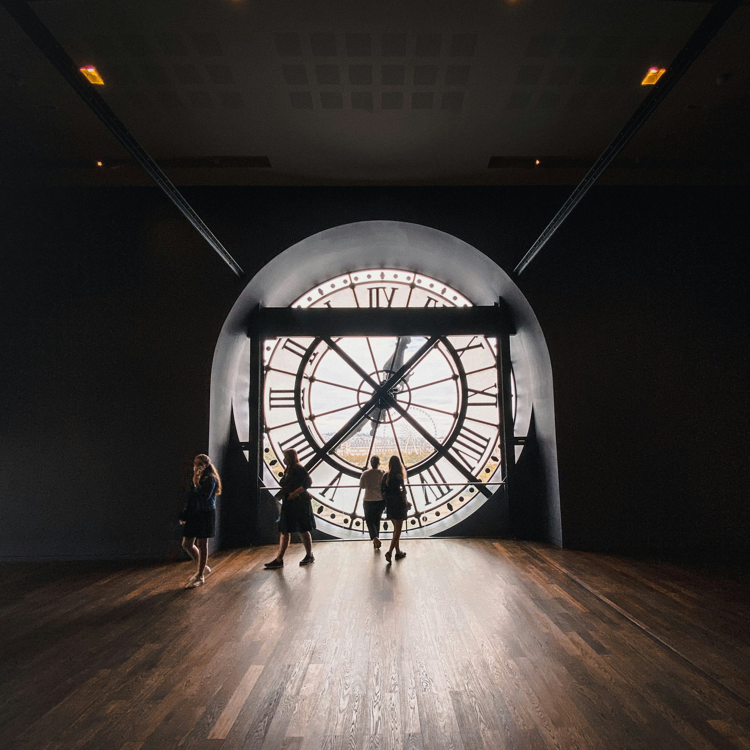 Four people standing and walking in front of a large clock face with Roman numerals inside a dark room.