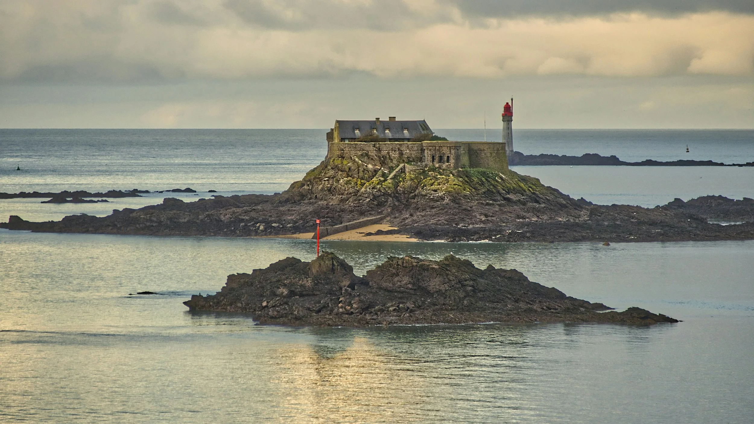 A lighthouse on a rocky island with an old building, surrounded by water and smaller rocks, under a cloudy sky.