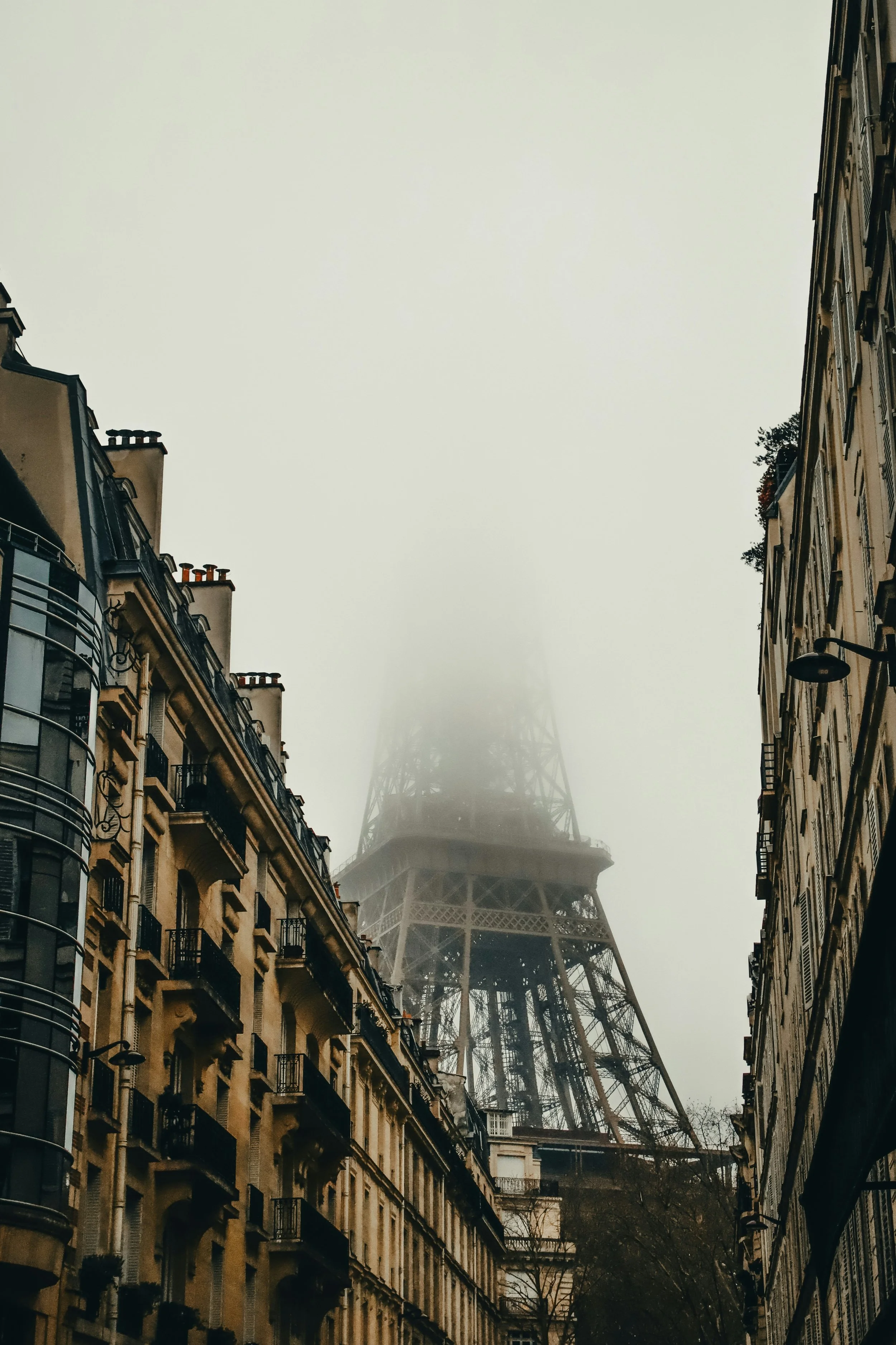 The Eiffel Tower partially obscured by fog, seen between two streets lined with traditional Parisian buildings.