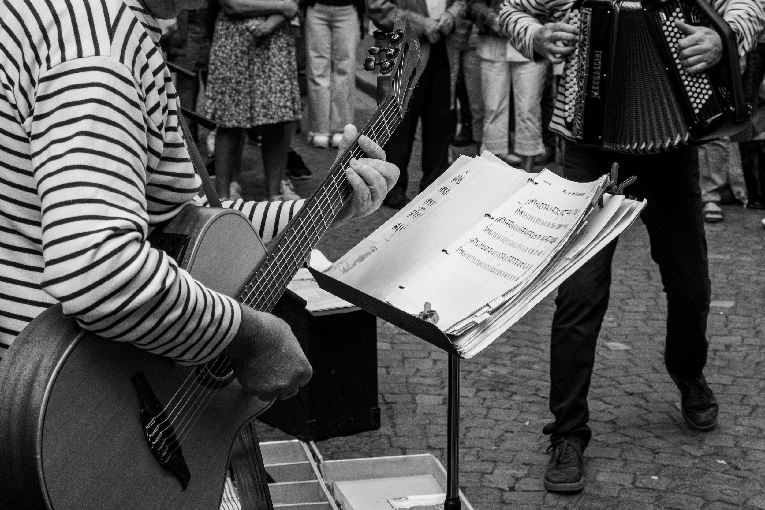 Street musician playing acoustic guitar and singing in front of a music stand with sheet music, crowd of people watching nearby, another musician playing an accordion.