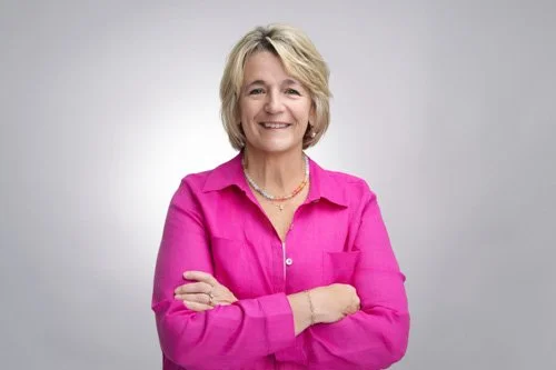 A woman with short blonde hair, wearing earrings, a brown zip-up jacket, and a crossbody bag, smiling outdoors near a black metal fence.