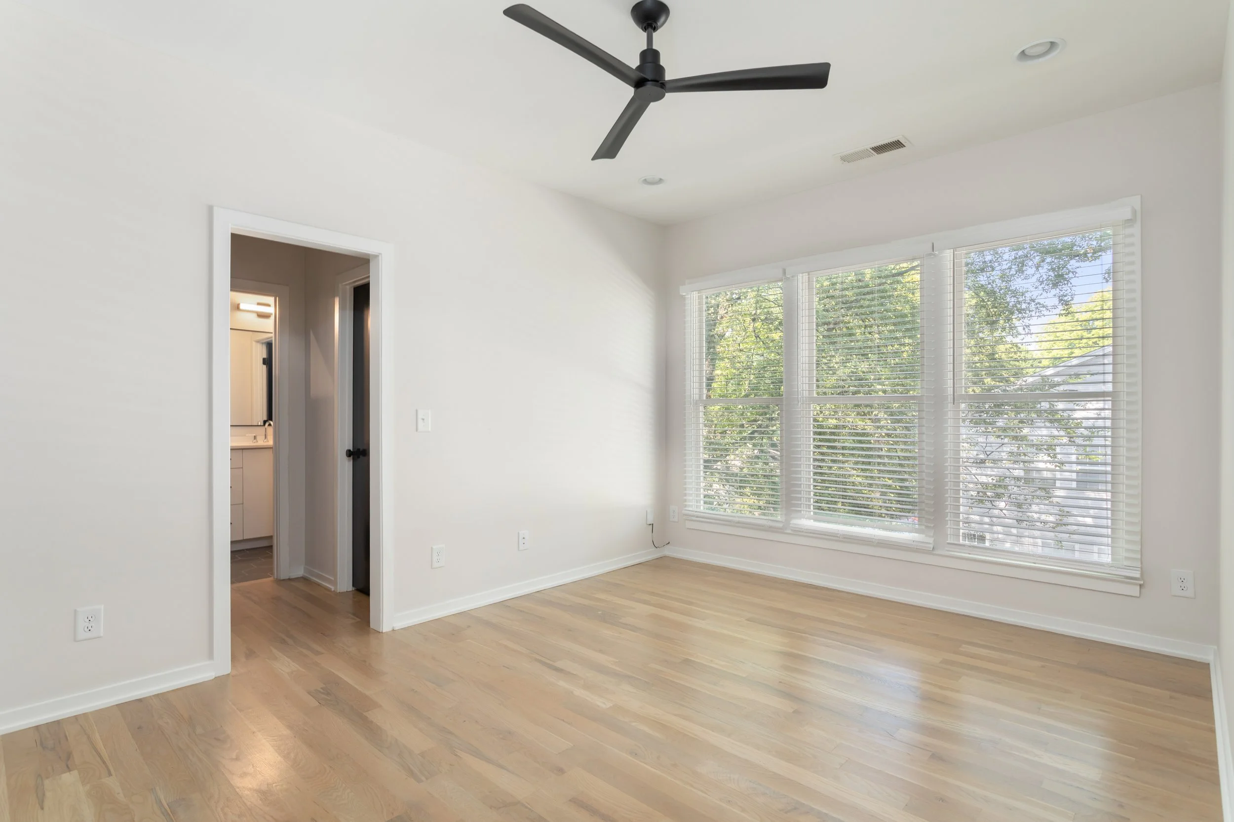 Bright empty room with hardwood floors, large windows with white blinds, and a modern black ceiling fan