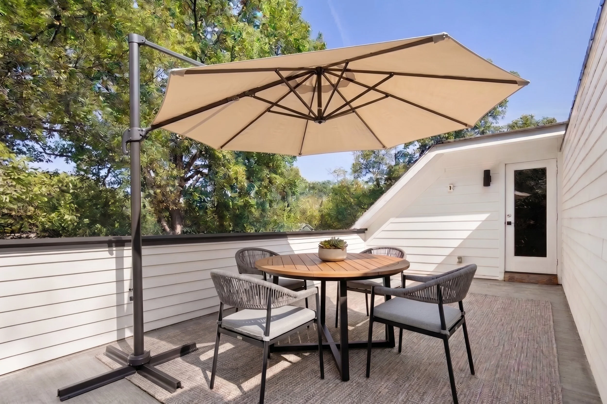 Outdoor patio dining set with a large beige umbrella, round wooden table, four chairs, and greenery beside a white house