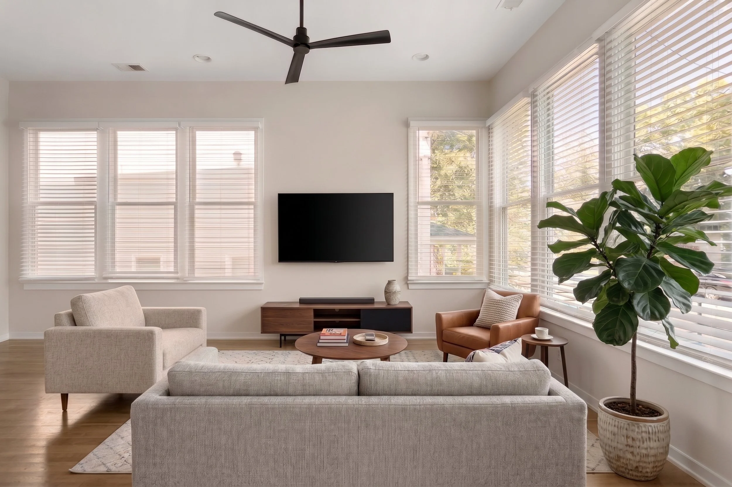 Bright modern living room with a wall-mounted TV, sectional sofa, armchairs, ceiling fan, large windows with blinds, coffee table, and potted fiddle-leaf fig tree