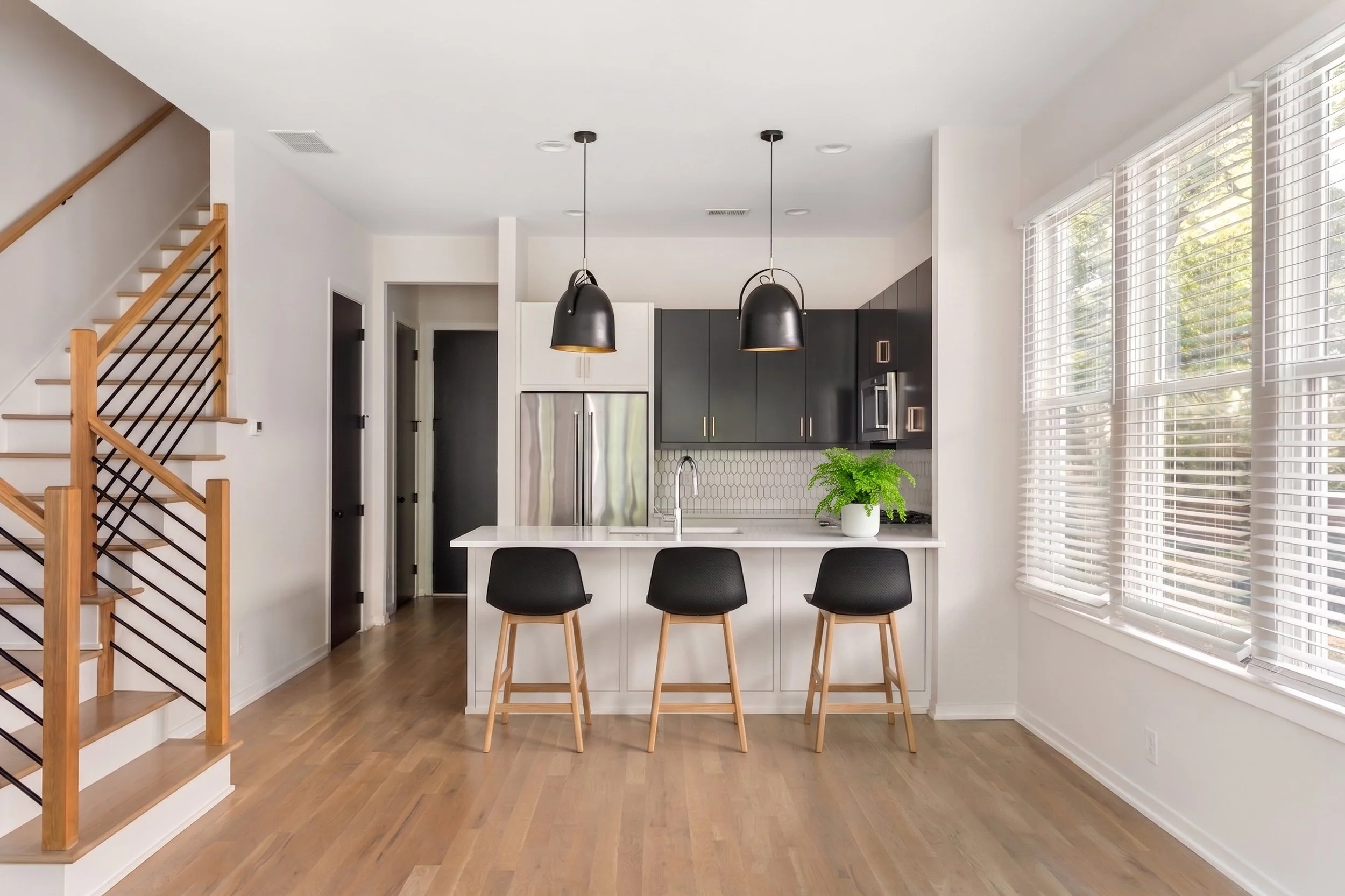 Modern open-plan kitchen with black cabinets, white island, pendant lights, three bar stools, wood staircase, and large windows with blinds