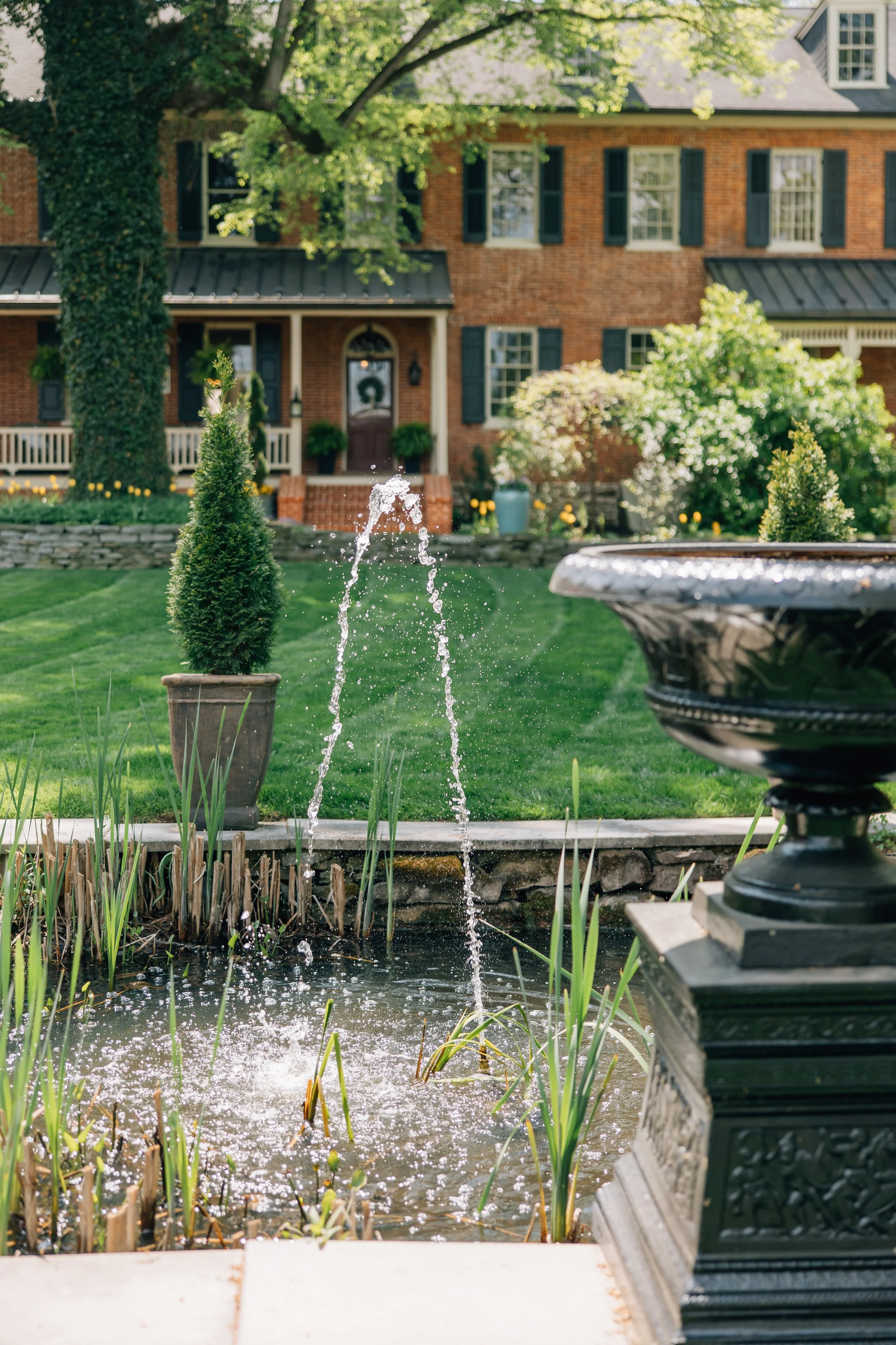 View of a pond with small water fountain, potted plants, and a house with brick exterior and green shutters in the background.