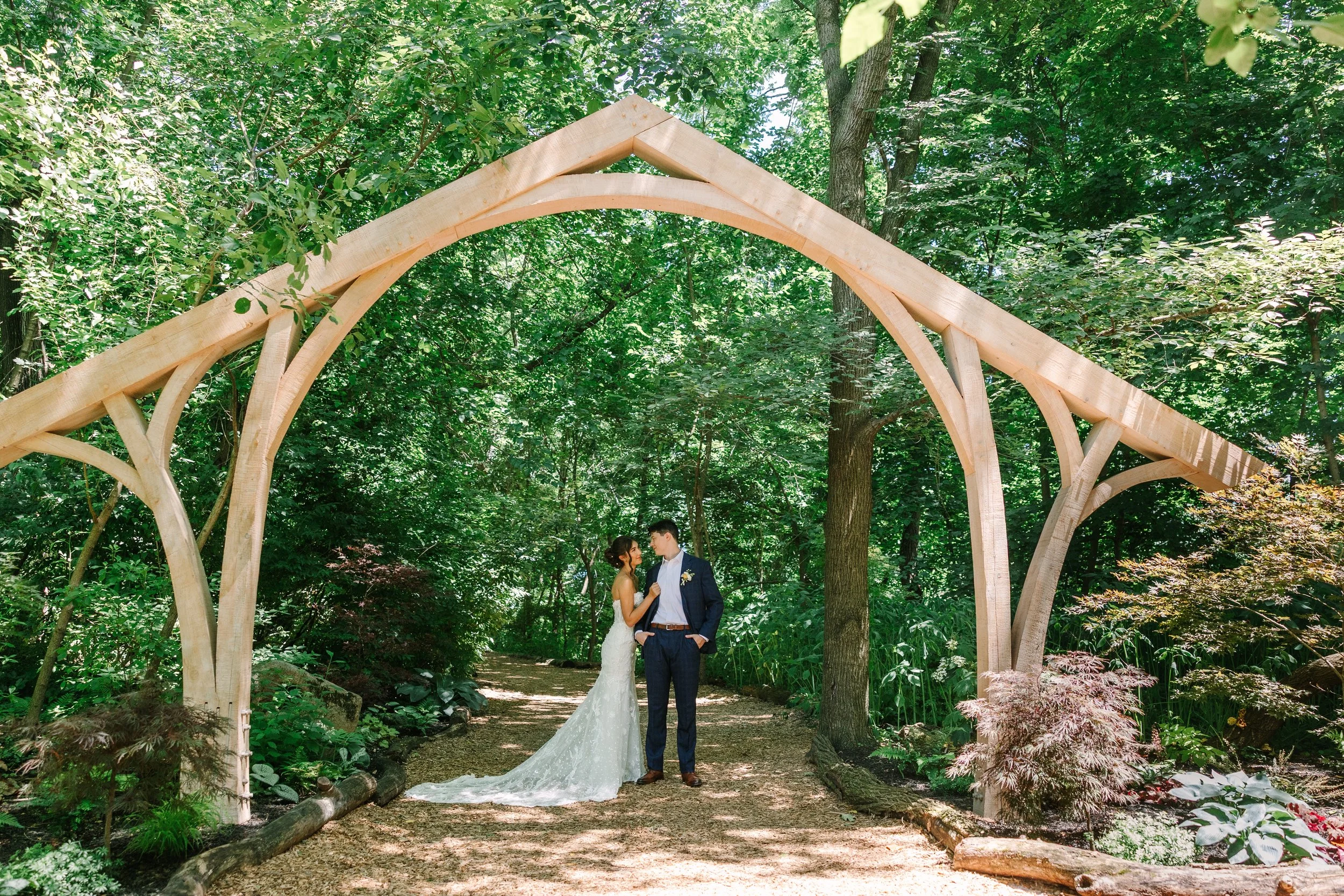 A bride and groom standing under a wooden arch structure in a lush green forest during a wedding ceremony.