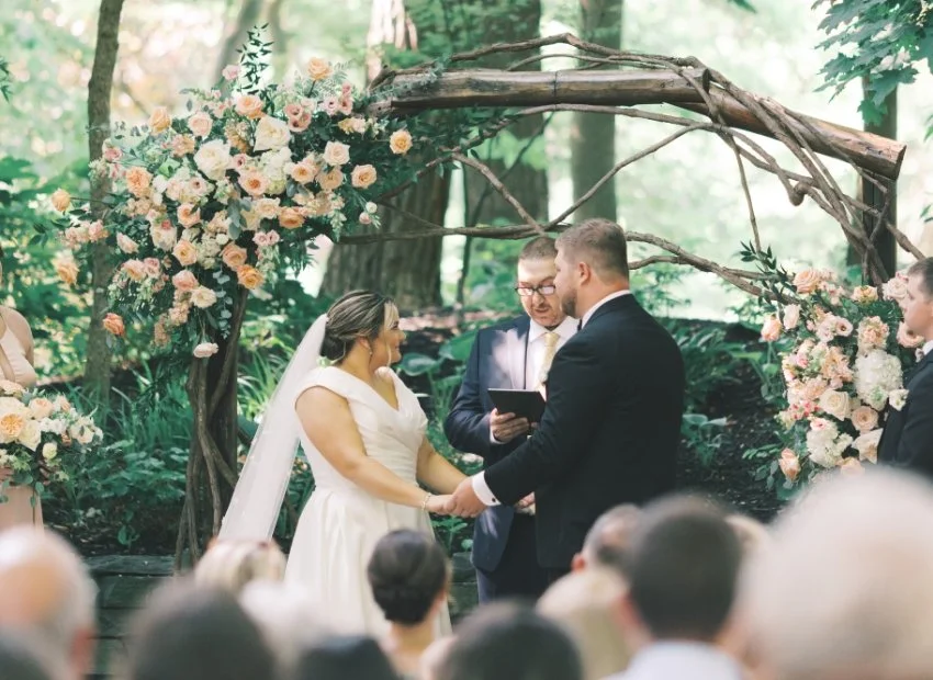 A couple gets married outdoors in a wooded area, with a floral arch behind them. The bride wears a white dress and veil, and the groom in a dark suit. An officiant reads from a tablet, and guests watch. Flowers are pink and white.