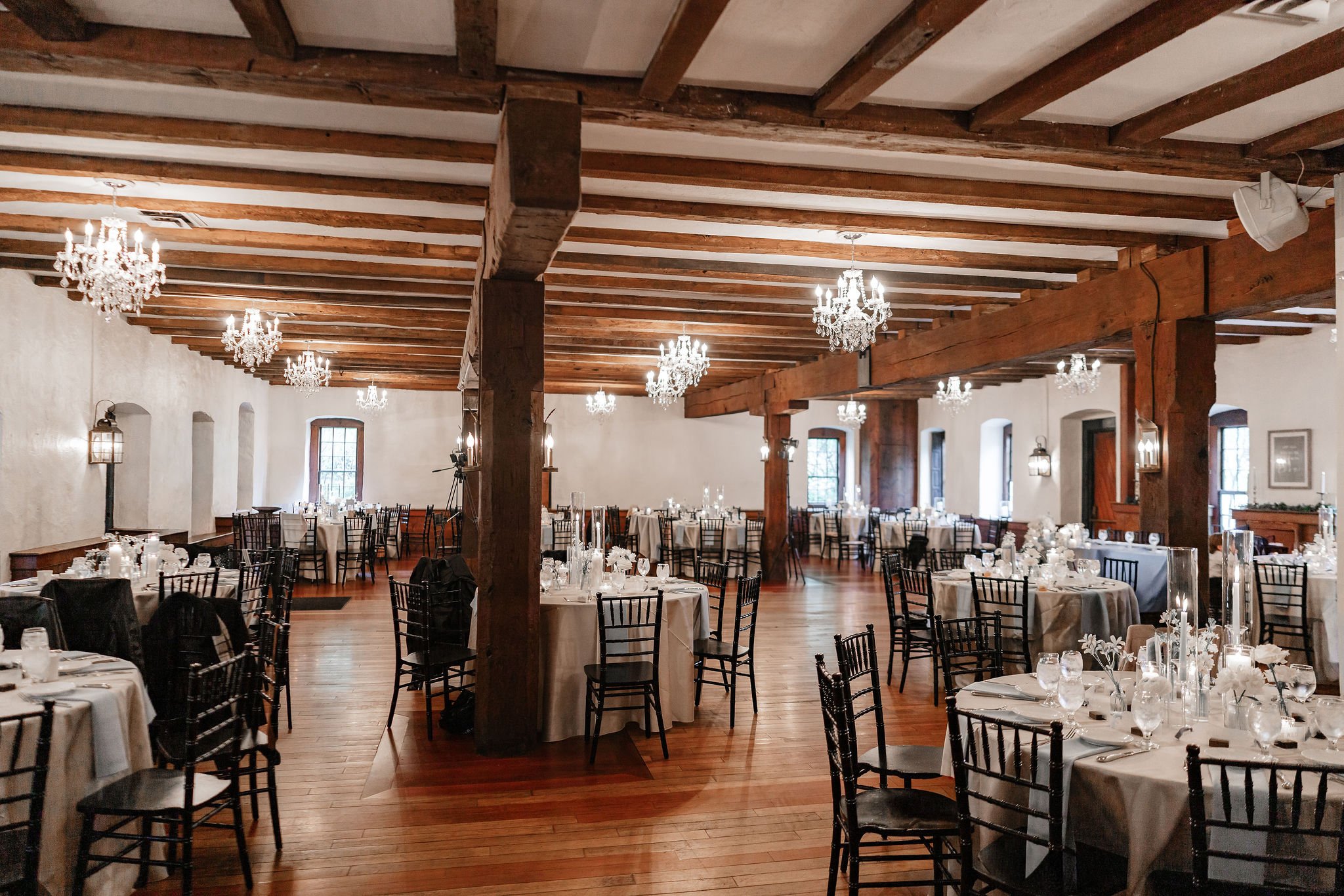 Interior of a banquet hall with wooden beams on the ceiling, crystal chandeliers, and round tables set with white tablecloths, glassware, and floral centerpieces.