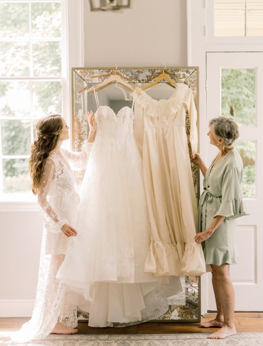 A young woman in a lace gown and an older woman in a pastel robe stand in front of a mirror, admiring two wedding dresses hanging on a decorative screen inside a bright room with large windows.