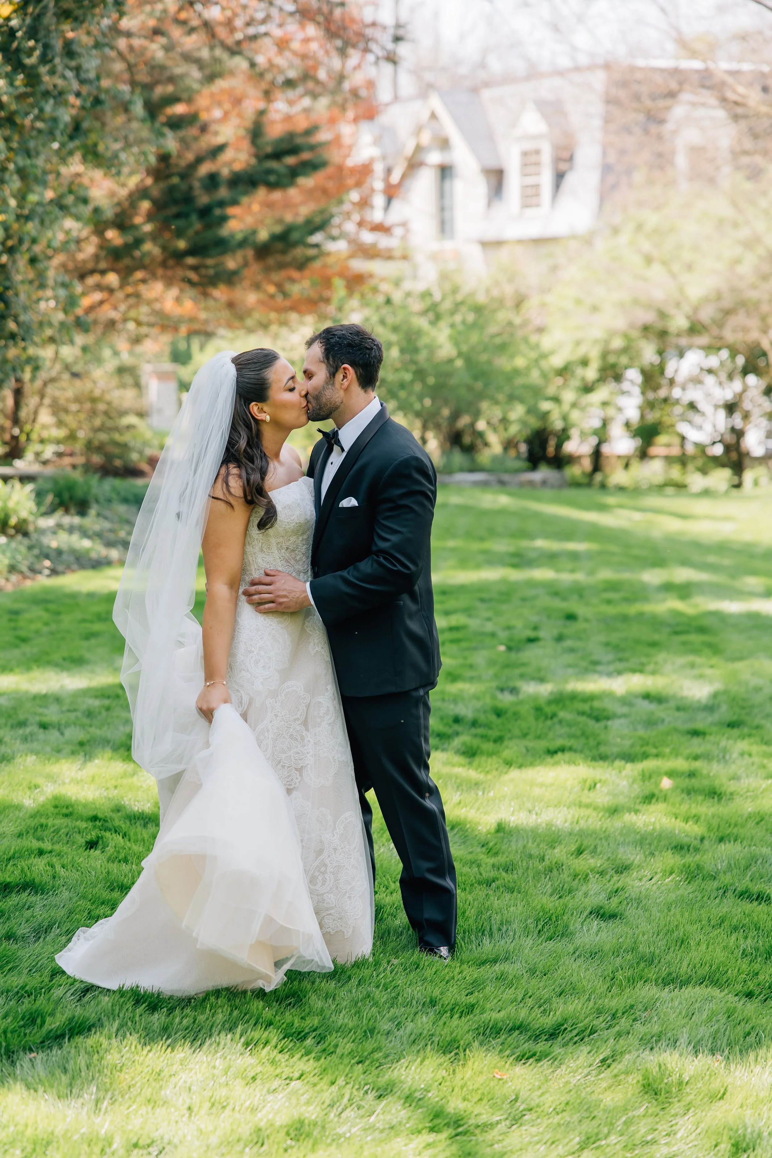 Bride and groom sharing a kiss outdoors on a sunny day, surrounded by green grass and trees with autumn-colored leaves, near an old house in the background.