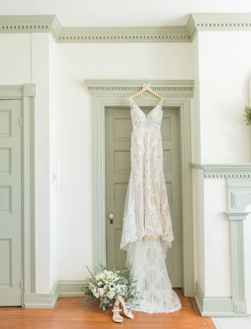 A wedding dress hanging on a hanger labeled "Bride" in front of a door, with a floral bouquet and a pair of white shoes on the floor.