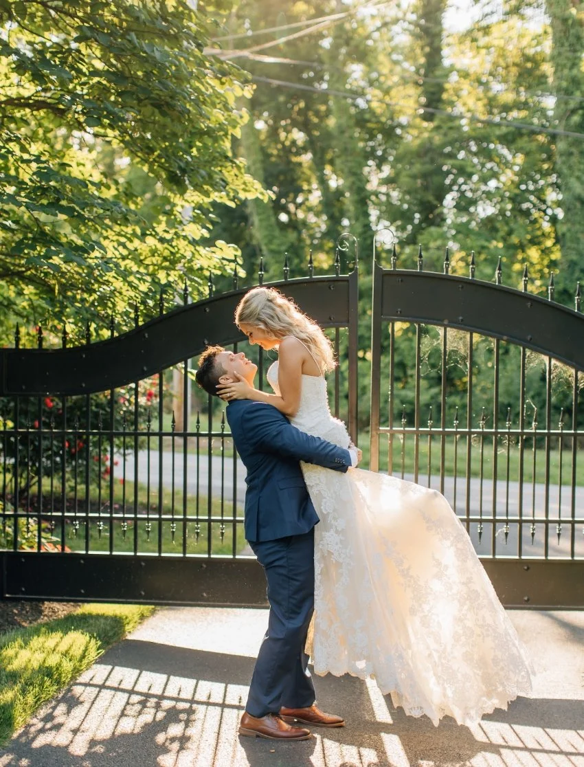 A couple in wedding attire embracing in front of a black wrought iron gate, with the groom lifting the bride, surrounded by green trees and sunlight.