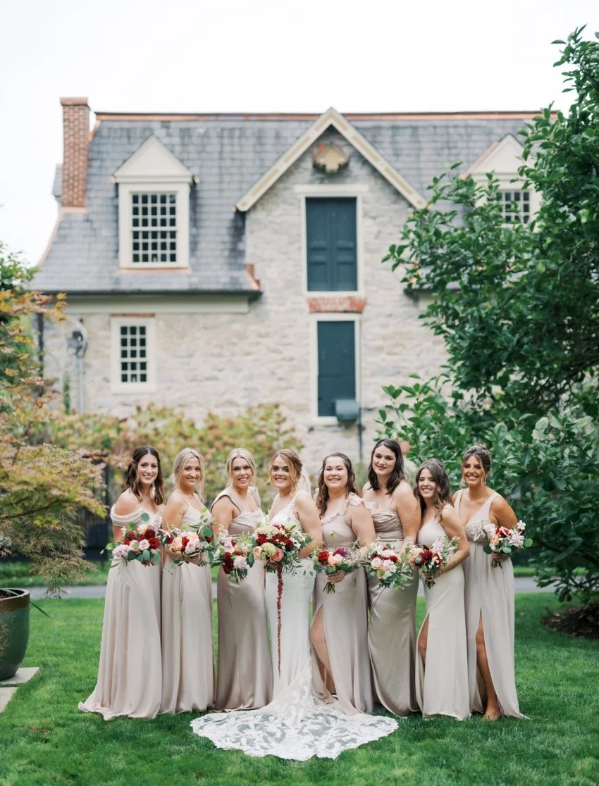 Group of eight women in bridesmaid dresses holding bouquets, standing on a lawn with a stone house in the background.